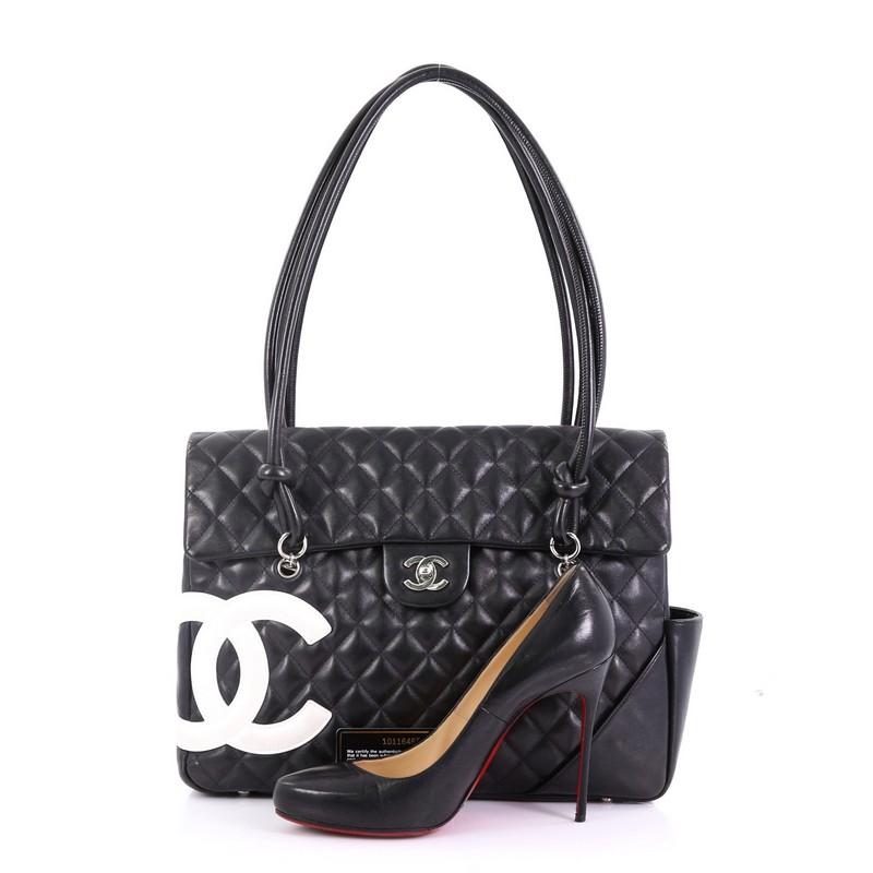 This Chanel Cambon Flap Tote Quilted Leather Large, crafted in black quilted leather, features tall handles with knotted ends, signature white contrast CC logo patch design, protective base studs, exterior side and back pockets, and silver-tone
