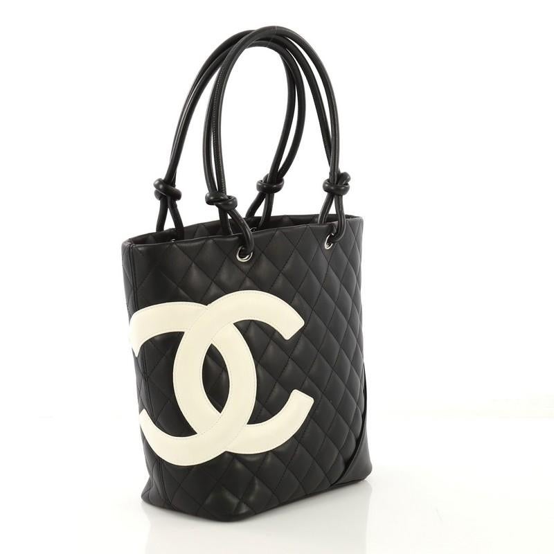 This Chanel Cambon Tote Quilted Leather Medium, crafted from black quilted leather, features white leather interlocking CC logo, dual rolled leather handles with knotted ends, exterior back pocket, and silver-tone hardware. Its zip closure opens to