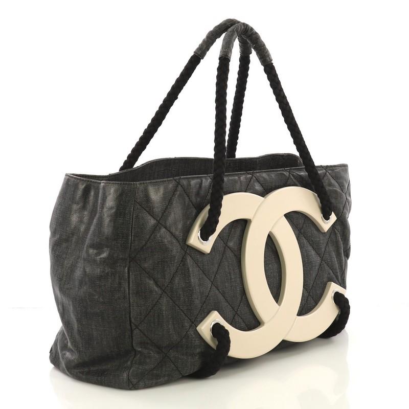 This Chanel CC Beach Tote Quilted Coated Canvas Large, crafted in black quilted coated canvas, features dual braided handles, large white wooden interlocking CC logo at the front, and silver-tone hardware. It opens to a black fabric interior with