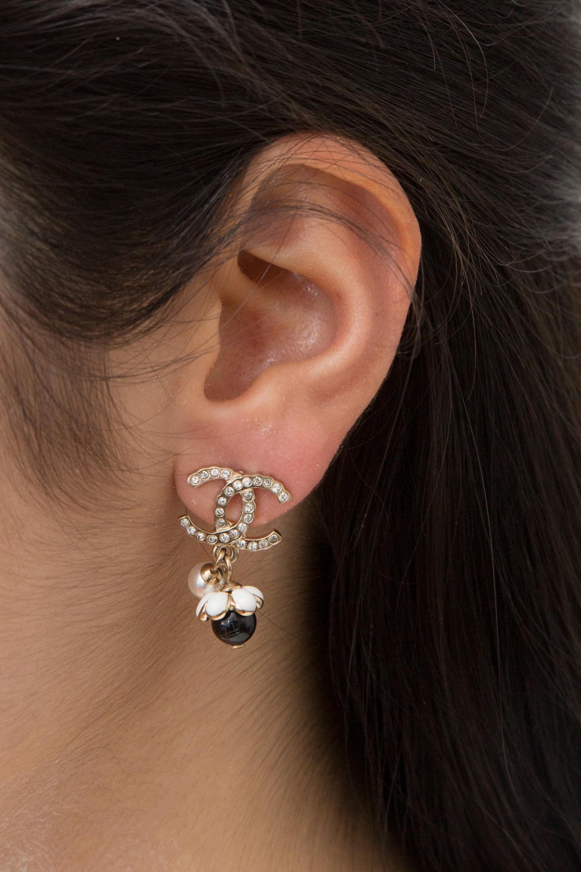 Chanel black and gold-tone “CC” pendant earrings featuring a central signature interlocking “CC” logo with crystal embellishment, and free faux-pearl, a flower with white and black pearls, and a back plaque pitted.  
They come as a pair. 
Circa