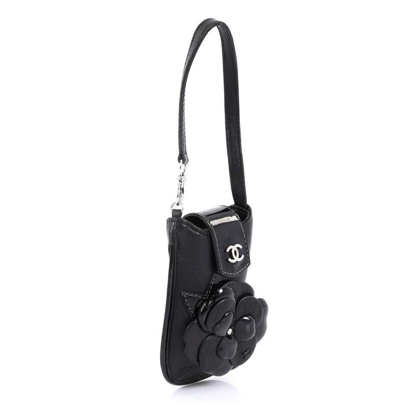 This Chanel CC Camellia Phone Holder Patent and Leather Embellished Lambskin, crafted in black patent and lambskin leather, features flat leather strap, camellia leather embellishment, and silver-tone hardware. Its snap closure opens to a black