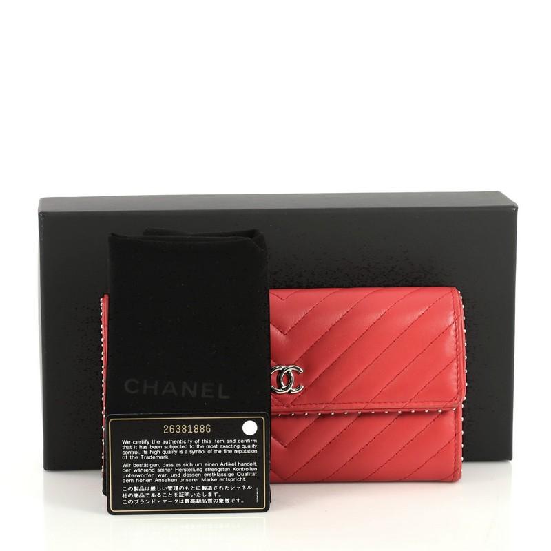 This Chanel CC Gusset Flap Wallet Chevron Lambskin with Studded Detail Long, crafted from red chevron lambskin leather, features interlocking CC logo, gusseted sides, stud trim and silver-tone hardware. Its snap button closure opens to a red leather