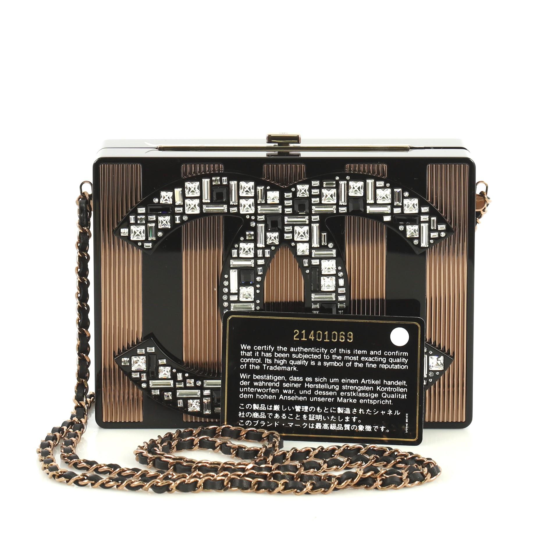 This Chanel CC Minaudiere Clutch Strass Embellished Plexiglass, crafted from black embellished plexiglass, features woven-in leather chain strap, CC logo embellished in strass, and copper-tone hardware. It opens to a black leather interior. Hologram