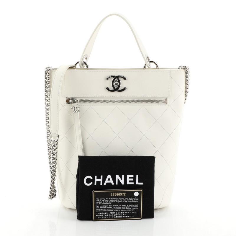 This Chanel CC Top Handle Bucket Bag Stitched Calfskin Small, crafted from white stitched calfskin leather, features a leather top handle, chain strap with leather pad, and silver-tone hardware. Its CC turn-lock closure opens to a neutral fabric