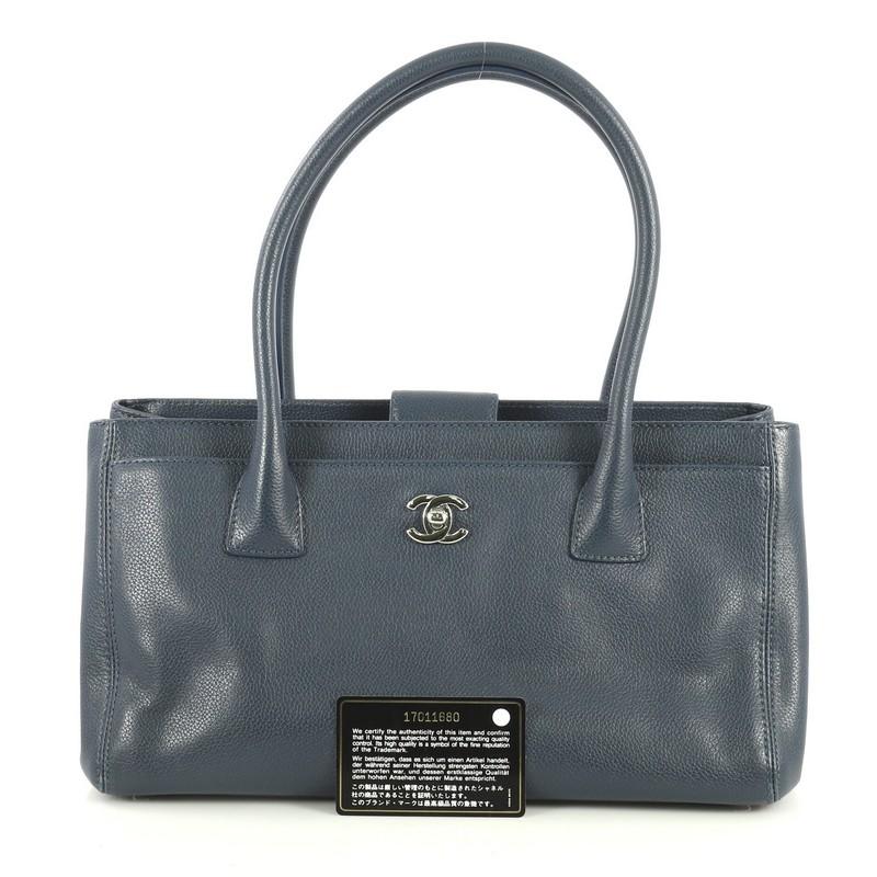 This Chanel Cerf Executive Tote Leather Small, crafted from blue leather, features dual rolled tall handles, front pocket with CC turn-lock closure, protective base studs, and silver-tone hardware. Its magnetic snap closure opens to a gray fabric