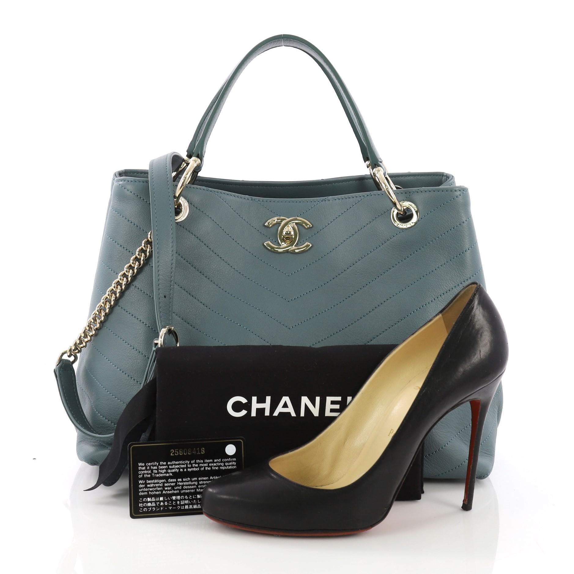 This Chanel Chevron Chic Shopping Tote Chevron Calfskin Small, crafted from teal chevron calfskin leather, features dual top handles, woven-in leather straps, protective base studs, and gold-tone hardware. Its CC turn-lock closure opens to a