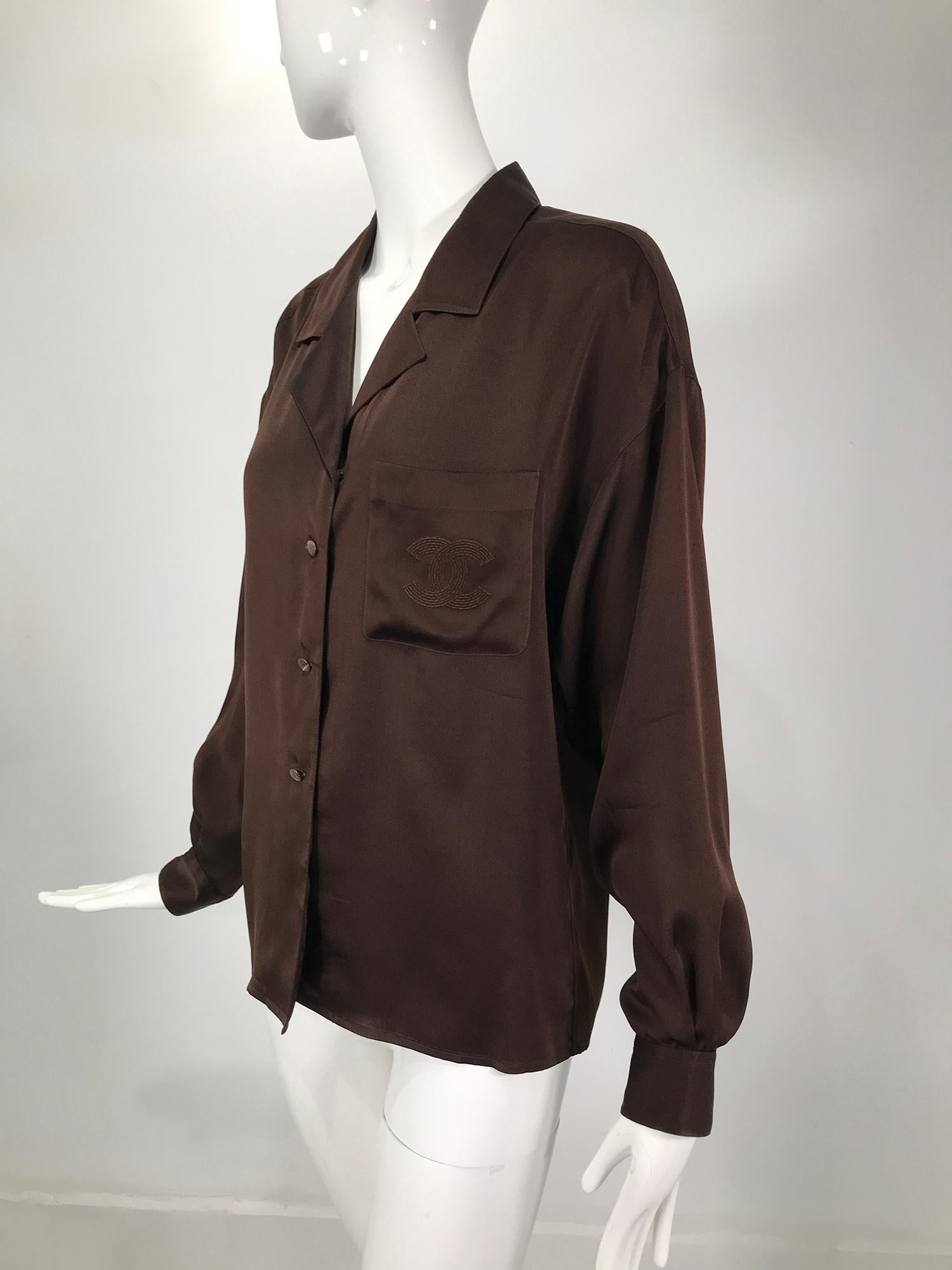 Chanel chocolate brown silk satin blouse with logo embroidered pocket. Long sleeve button front blouse with dropped shoulder yoke at front and back, wing collar, single left front breast logo embroidered pocket. The blouse closes at the front with