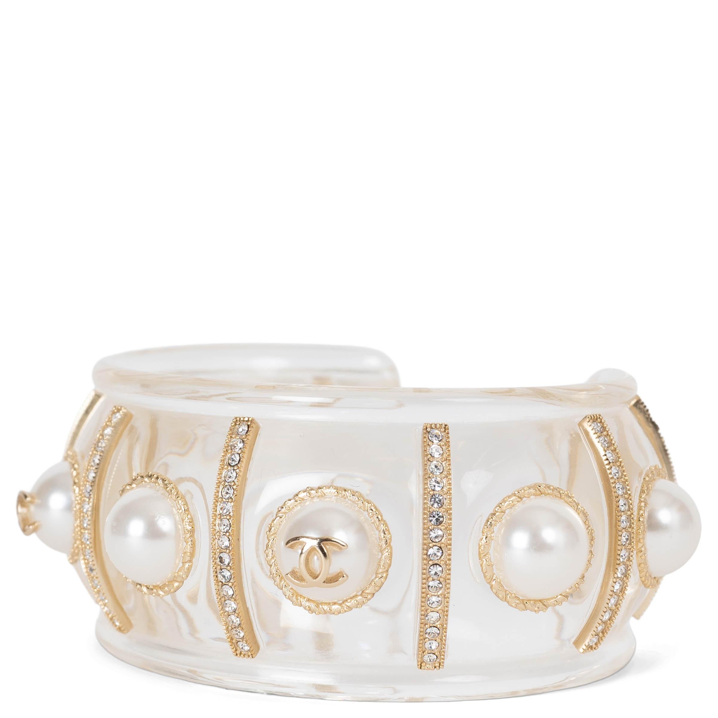 Este brazalete 100% auténtico de Chanel está fabricado en lucita transparente y metal dorado, y desprende una elegancia moderna con un toque característico. El diseño está adornado con tiras alternas de cristales transparentes y cabujones de perlas