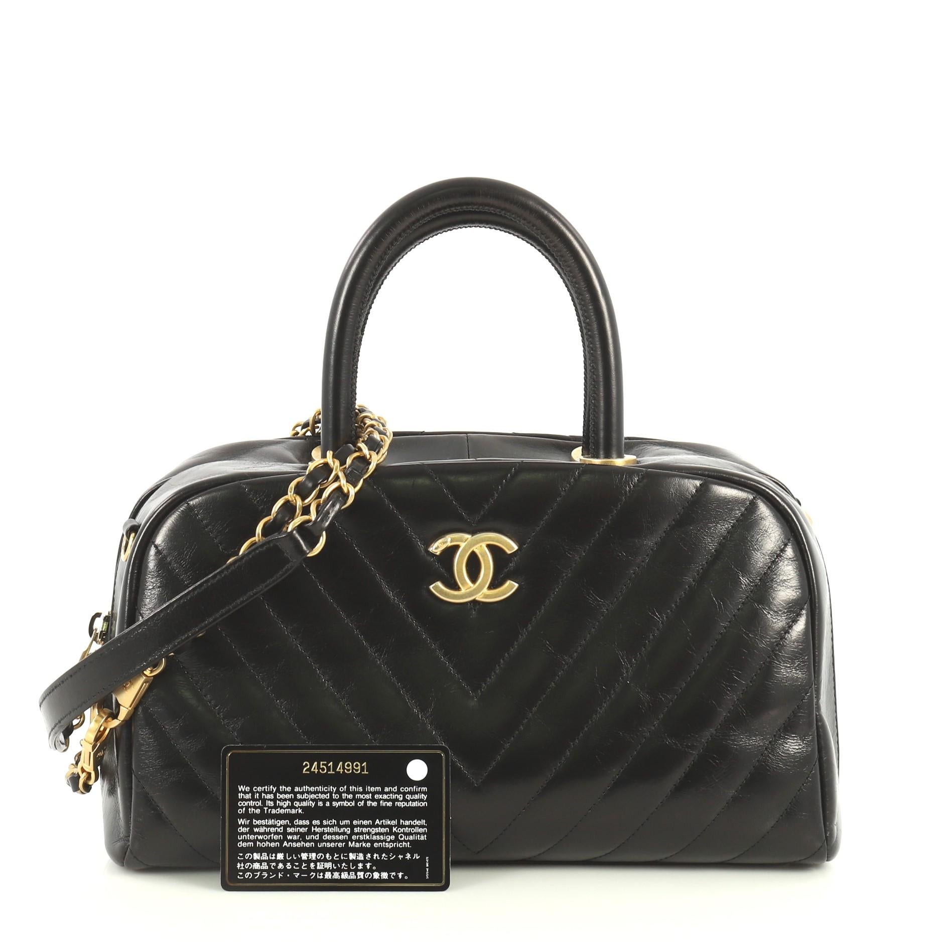 This Chanel Coco Bowling Bag Chevron Glazed Calfskin Small, crafted from black chevron glazed calfskin leather, features dual rolled handles and aged gold-tone hardware. Its zip closure opens to a red fabric interior with zip and slip pockets.