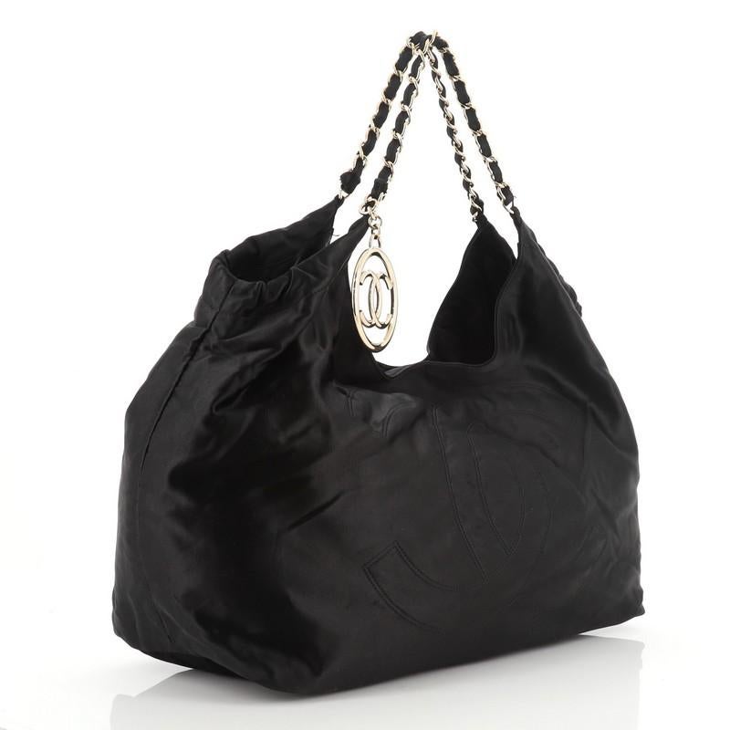 This Chanel Coco Cabas Satin Large, crafted from black satin, features an oversized stitched CC logo, woven-in satin chain straps, and gold-tone hardware. Its magnetic snap closure opens to a black fabric interior with side zip and slip pockets.