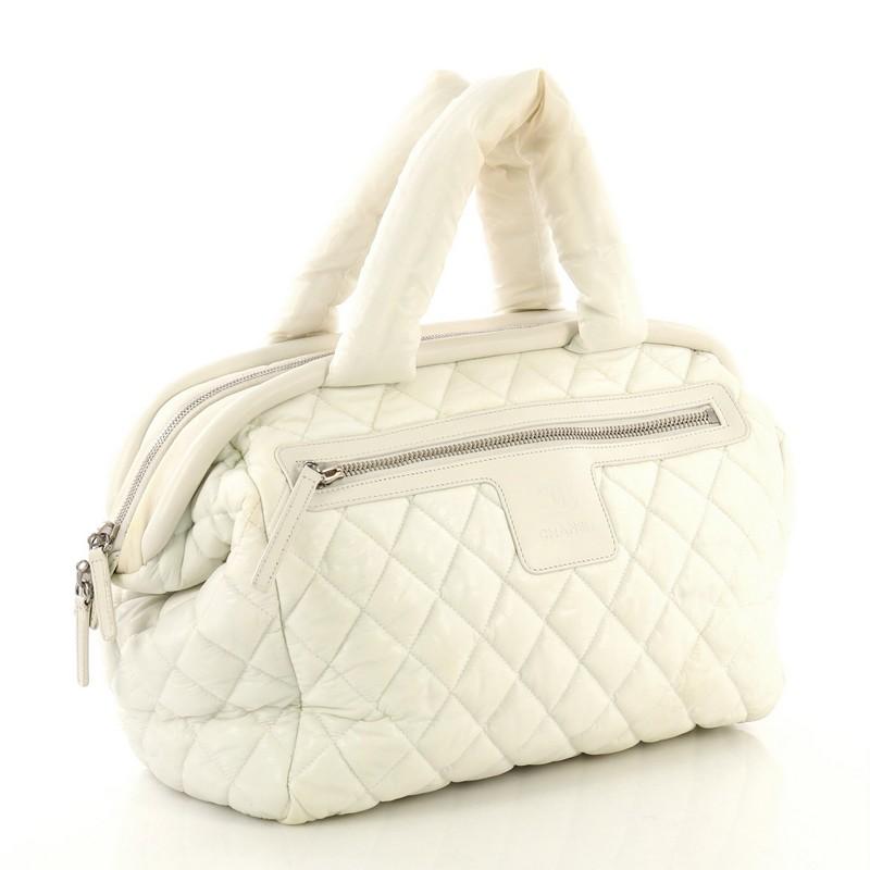 This Chanel Coco Cocoon Bowling Bag Quilted Nylon Medium, crafted from white quilted nylon, features dual nylon handles, exterior front zip pocket, and silver-tone hardware. Its zip closure opens to a blue nylon interior with zip pocket. Hologram