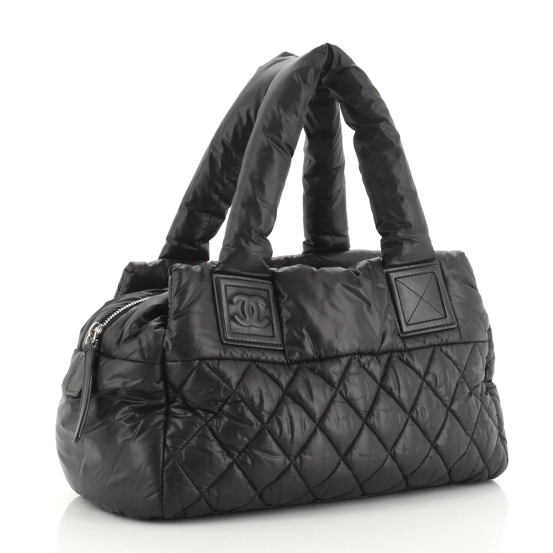 This Chanel Coco Cocoon Bowling Bag Quilted Nylon Small, crafted from black quilted nylon, features dual top handles, exterior front zip pocket, and silver-tone hardware. Its zip closure opens to a red nylon and fabric interior with zip pocket.