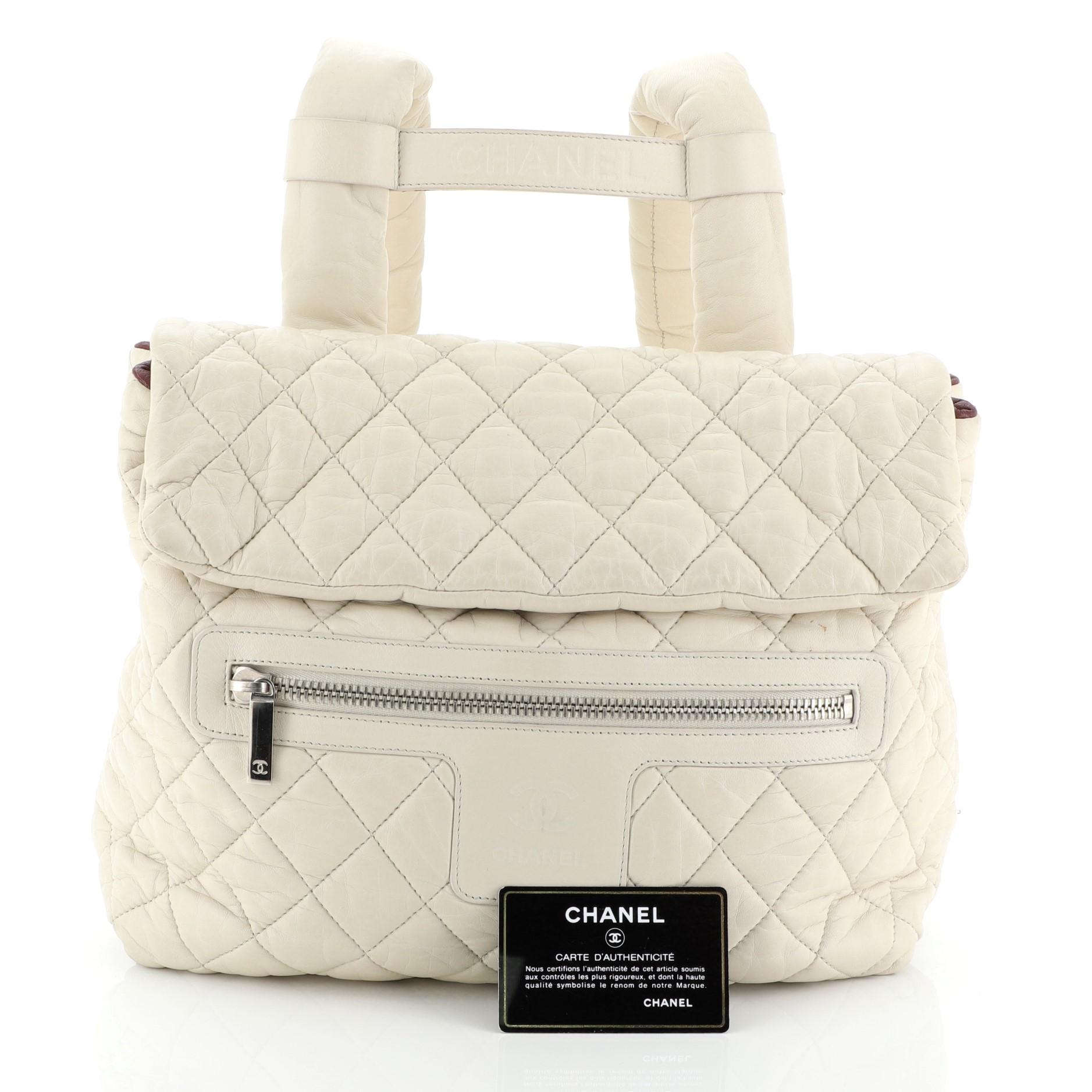This Chanel Coco Cocoon Flap Backpack Quilted Nylon, crafted in neutral quilted nylon, features dual backpack straps, leather trim, exterior zip pocket, front flap, and silver-tone hardware. Its magnetic snap closure opens to a red nylon interior