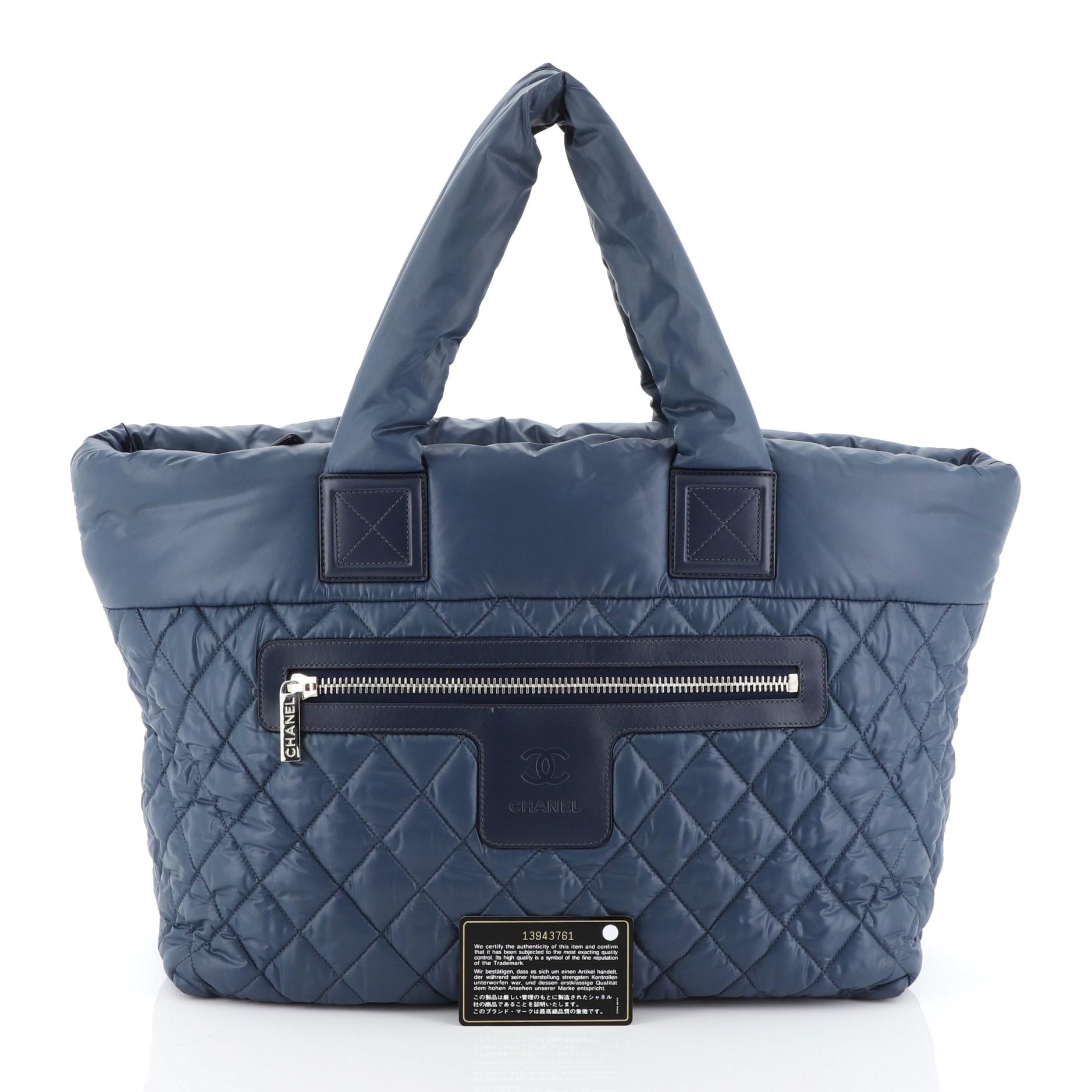 This Chanel Coco Cocoon Zipped Tote Quilted Nylon Medium, crafted from blue quilted nylon, features padded top handles, exterior zip pocket, and silver-tone hardware. Its zip closure opens to a black nylon interior with zip pocket. Hologram sticker