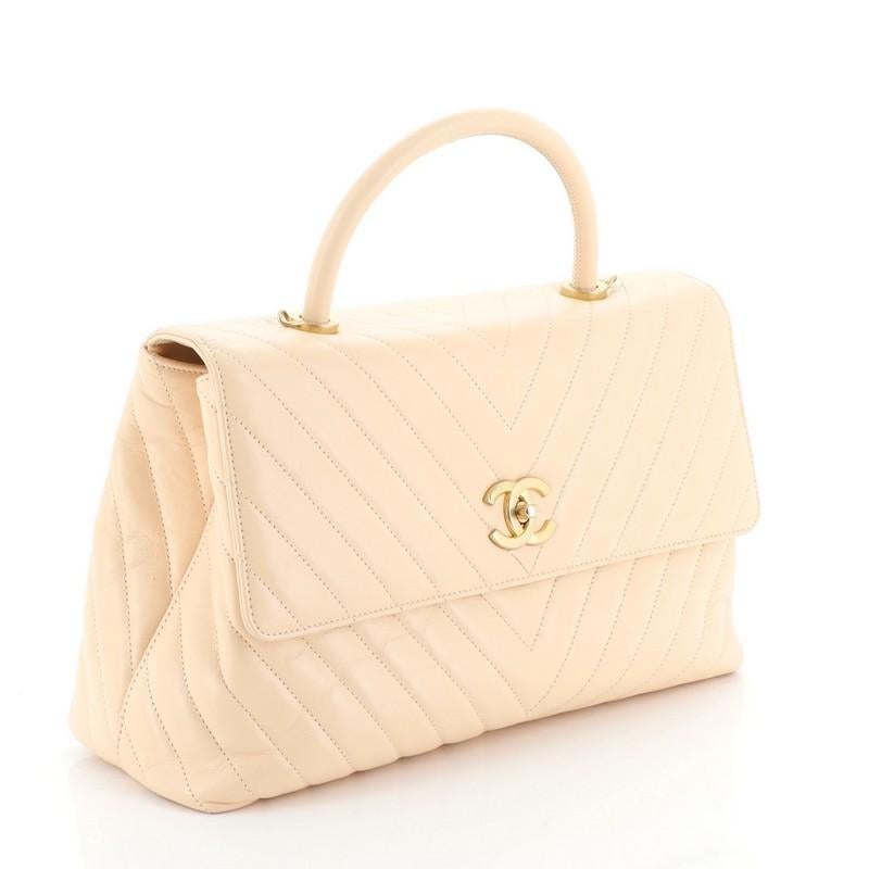 This Chanel Coco Top Handle Bag Chevron Calfskin Large, crafted in neutral chevron calfskin leather, features a single loop leather handle, exterior back slip pocket, and gold-tone hardware. Its CC turn-lock closure opens to neutral fabric interior