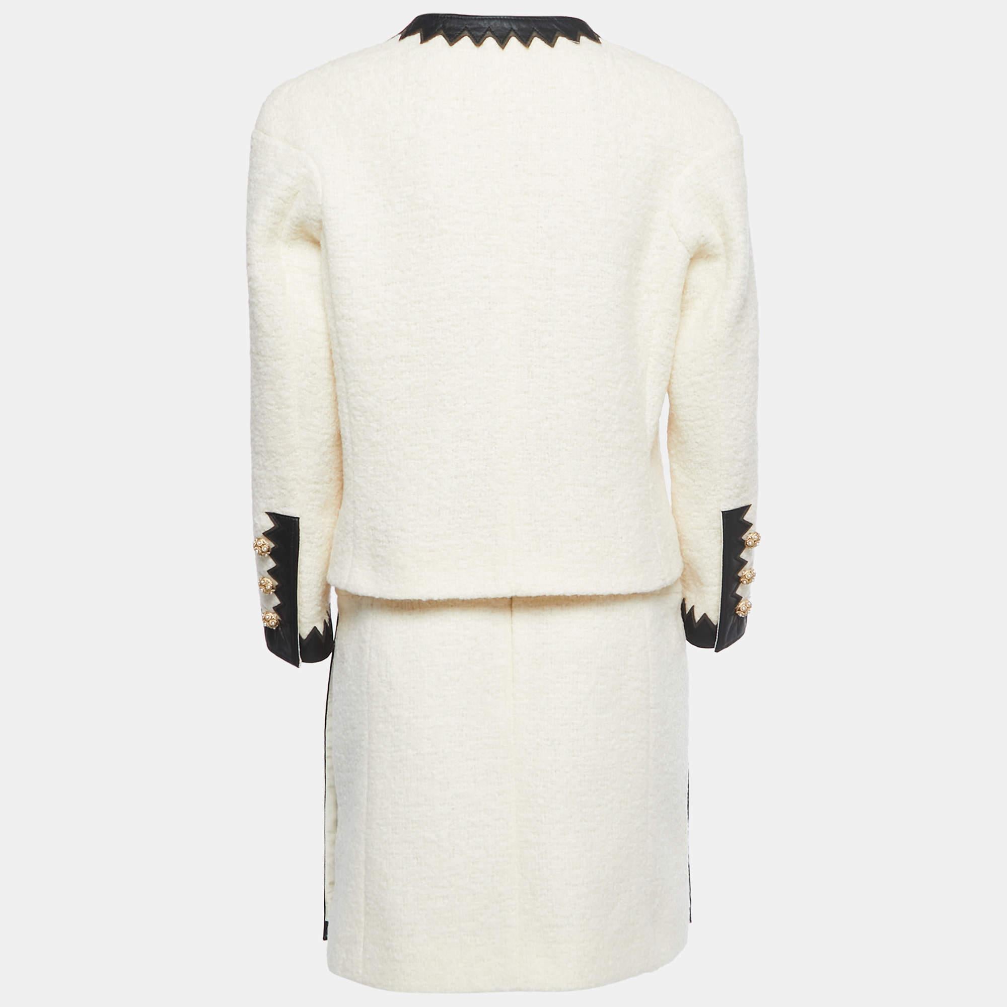 Le tailleur Chanel respire l'élégance avec son tissu en laine bouclée de couleur crème. Les bordures en cuir ajoutent une touche de luxe en rehaussant les revers et les poches de la veste. Un ensemble sophistiqué qui allie le style classique de