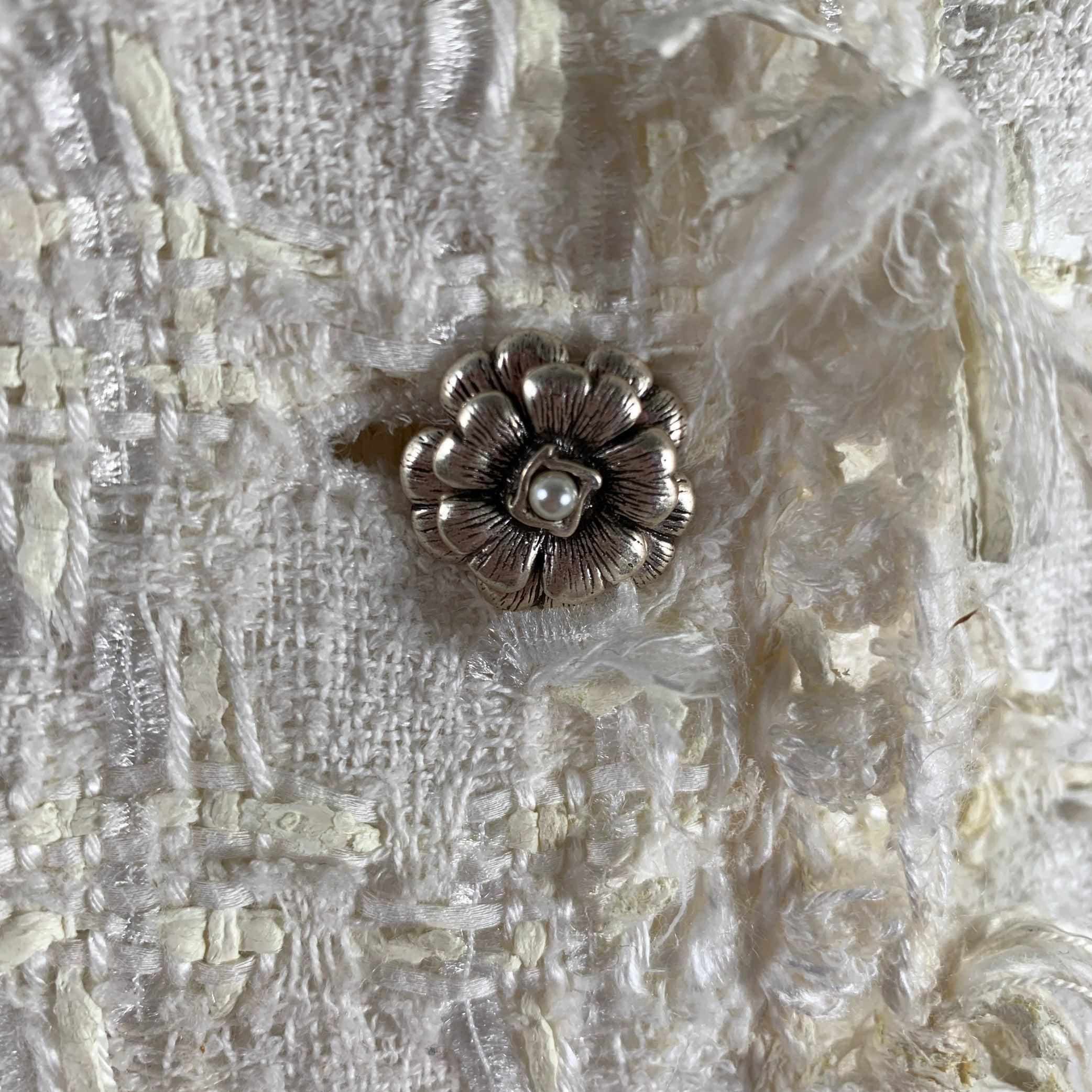 CHANEL jacket comes in a cream boucle cotton blend with a full monogram liner featuring a notch lapel, camelia pin detail, silver tone buttons, and a three button closure. Made in France. Very Good Pre-Owned Condition. Small mark at sleeve.
