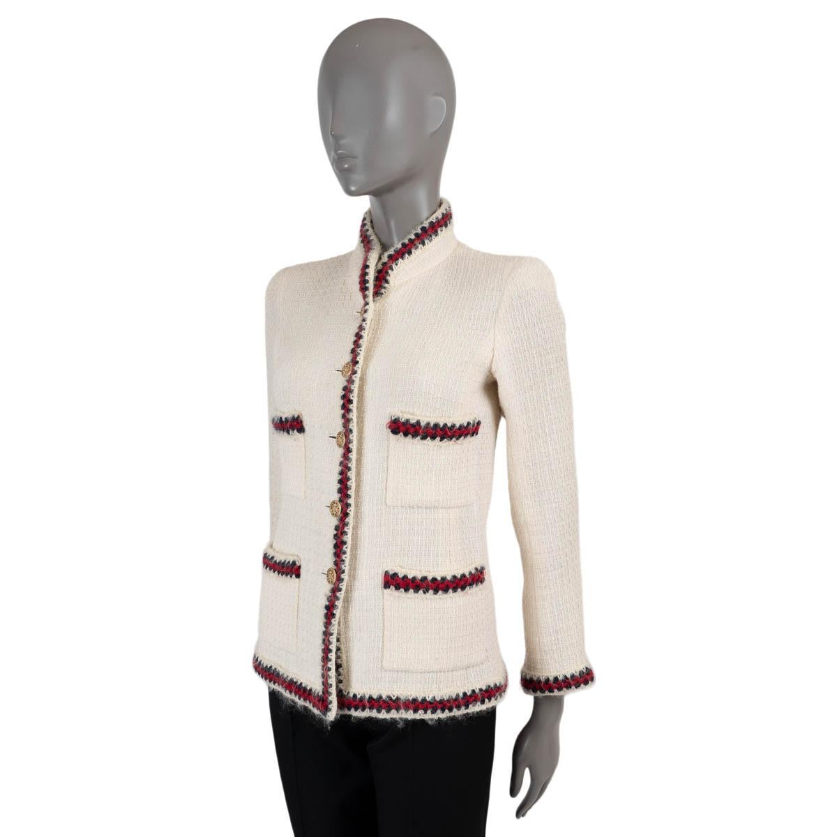100% authentische Chanel Tweedjacke aus ecrufarbener Wolle (100%). Mit rotem und dunkelblauem Häkelbesatz, Stehkragen und vier Taschen auf der Vorderseite. Wird mit goldfarbenen Knöpfen auf der Vorderseite geschlossen und ist mit Seide (mit 14%