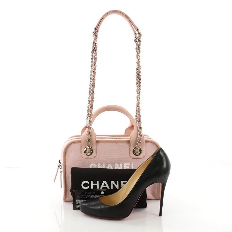 This Chanel Deauville Bowling Bag Canvas Small, crafted in pink canvas, features dual rolled top leather handles, dual woven-in chain straps with leather pads, printed Chanel logo and Parisian store address, and silver-tone hardware. Its zip closure