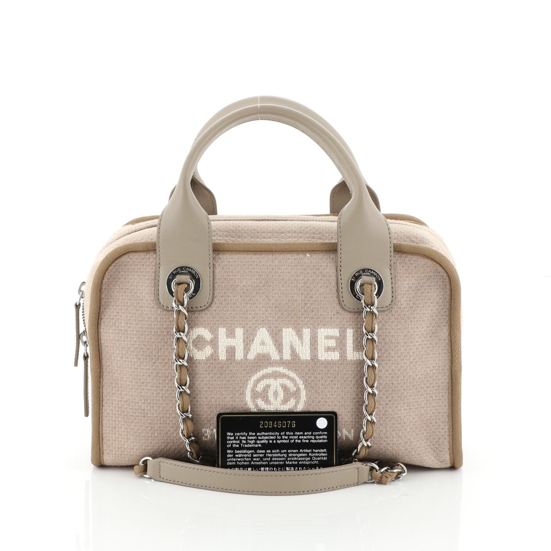 This Chanel Deauville Bowling Bag Canvas Small, crafted in neutral canvas, features dual rolled leather top handles, dual woven-in chain straps with leather pads, Chanel logo and Parisian store address on front, and silver-tone hardware. Its zip