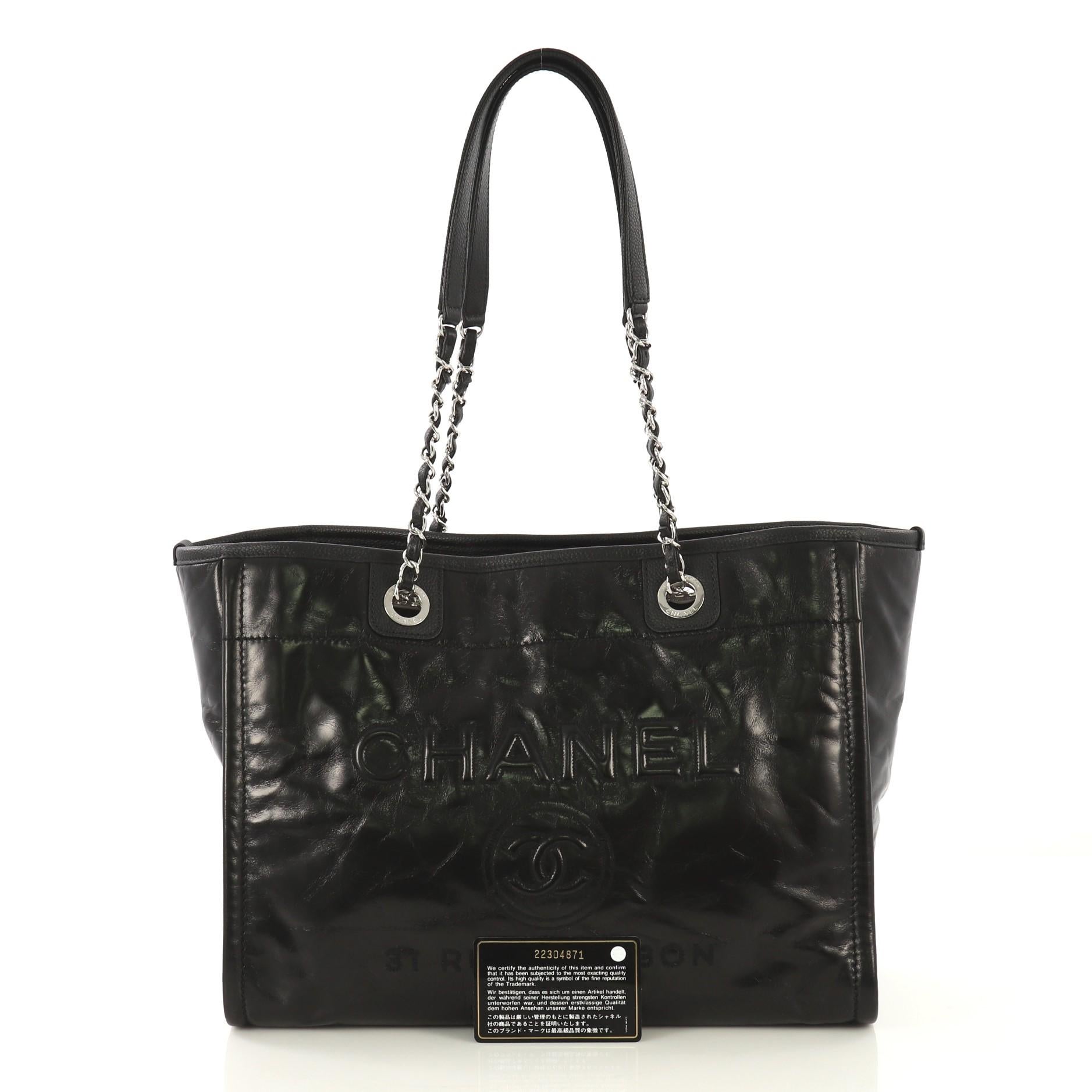 This Chanel Deauville Tote Glazed Calfskin Small, crafted in black glazed calfskin leather, features woven-in chain link straps, CC logo with Chanel's famous Parisian store address, quilted exterior back pocket, and silver-tone hardware. Its