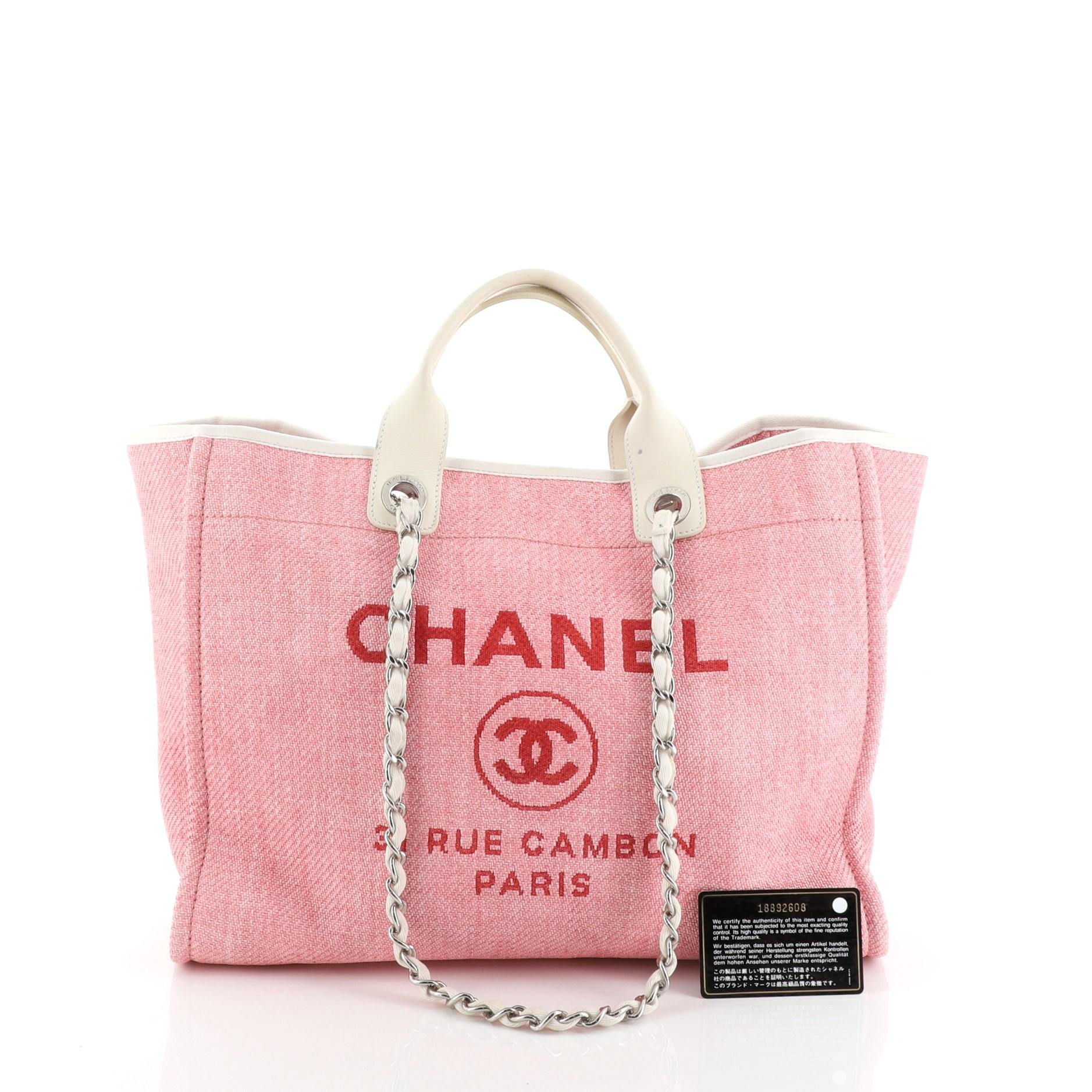This Chanel Deauville Tote Raffia Large, crafted from pink raffia, features dual leather handles, woven-in canvas chain straps, and silver-tone hardware. Its magnetic snap closure opens to a pink fabric interior with slip and zip pockets. Hologram