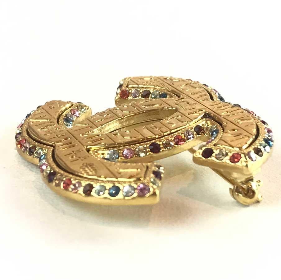 The brooch comes from Maison CHANEL and more particularly from the Métiers d'art Paris-New York Collection on the theme of Egypt. It represents the CC in gold-plated metal, which is set on all sides with multicolored rhinestones. Are engraved