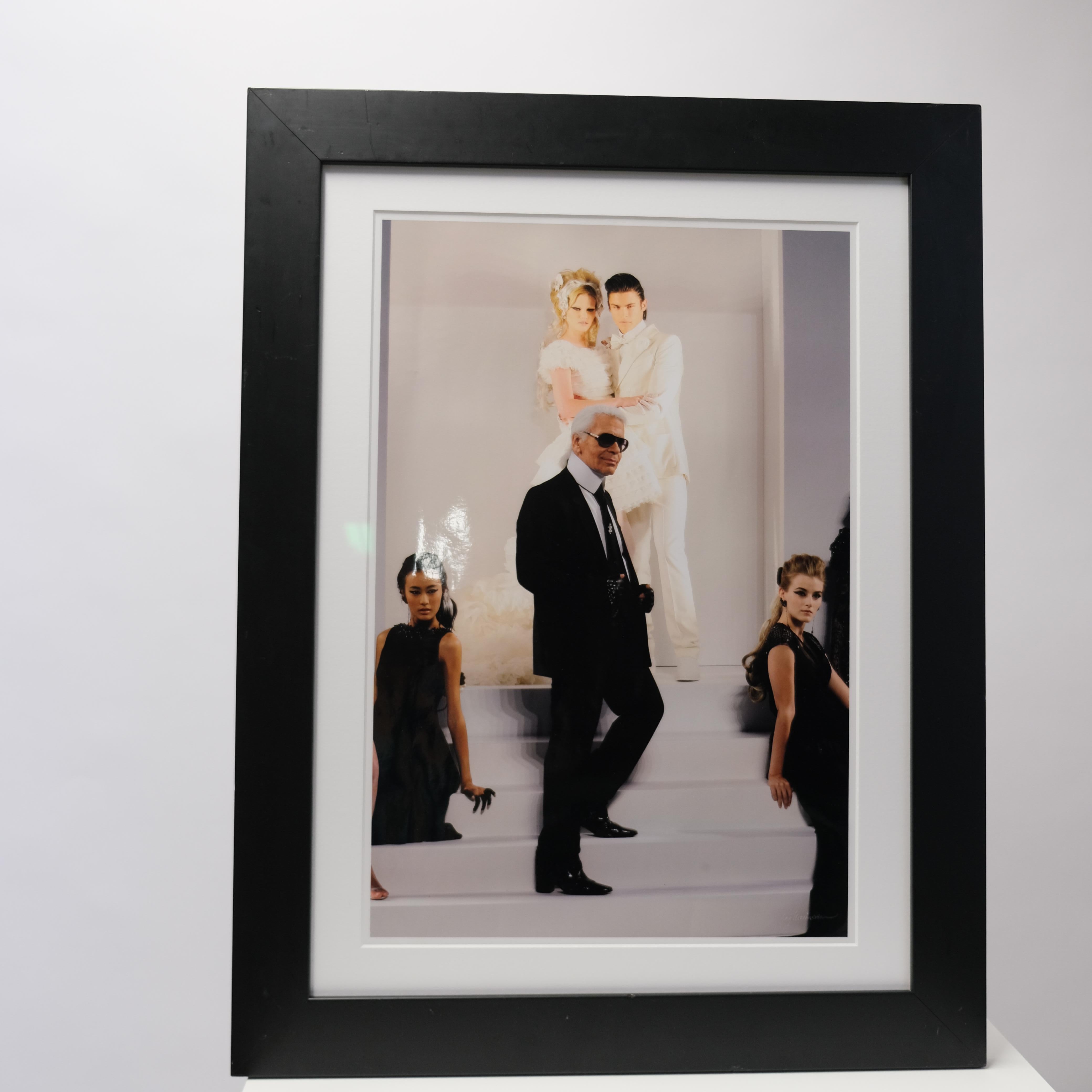 The photograph captures the legendary Karl Lagerfeld alongside Chanel models in a striking composition that embodies the essence of haute couture. Dressed in his signature black suit and sunglasses, Lagerfeld stands at the center, surrounded by