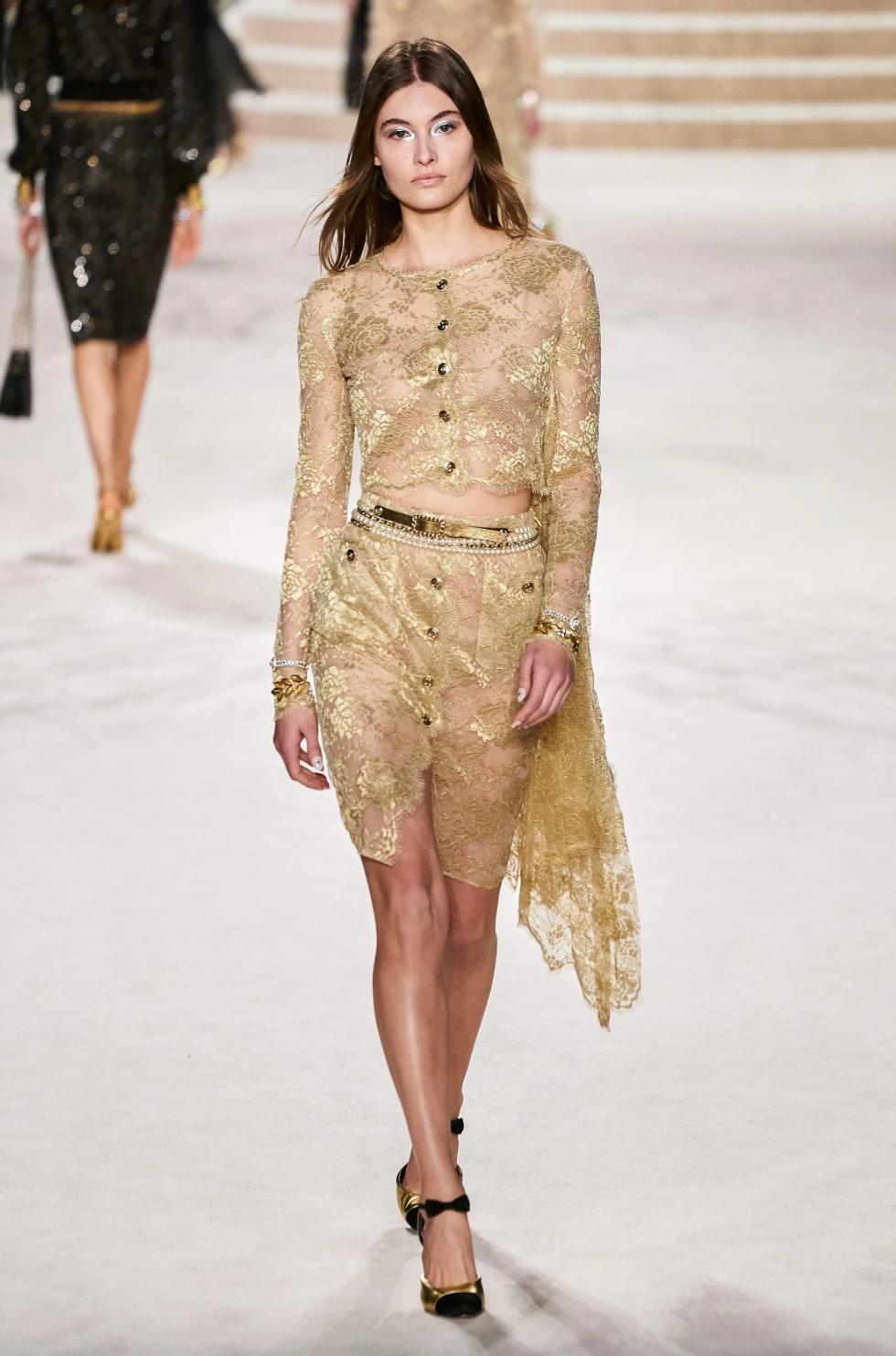 This exquisite Chanel two-piece suit from the Fall 2020 Métiers d'Art collection embodies elegance and sophistication with its intricate gold lace design. The ensemble features a semi-sheer, long-sleeve top and a matching pencil skirt, both