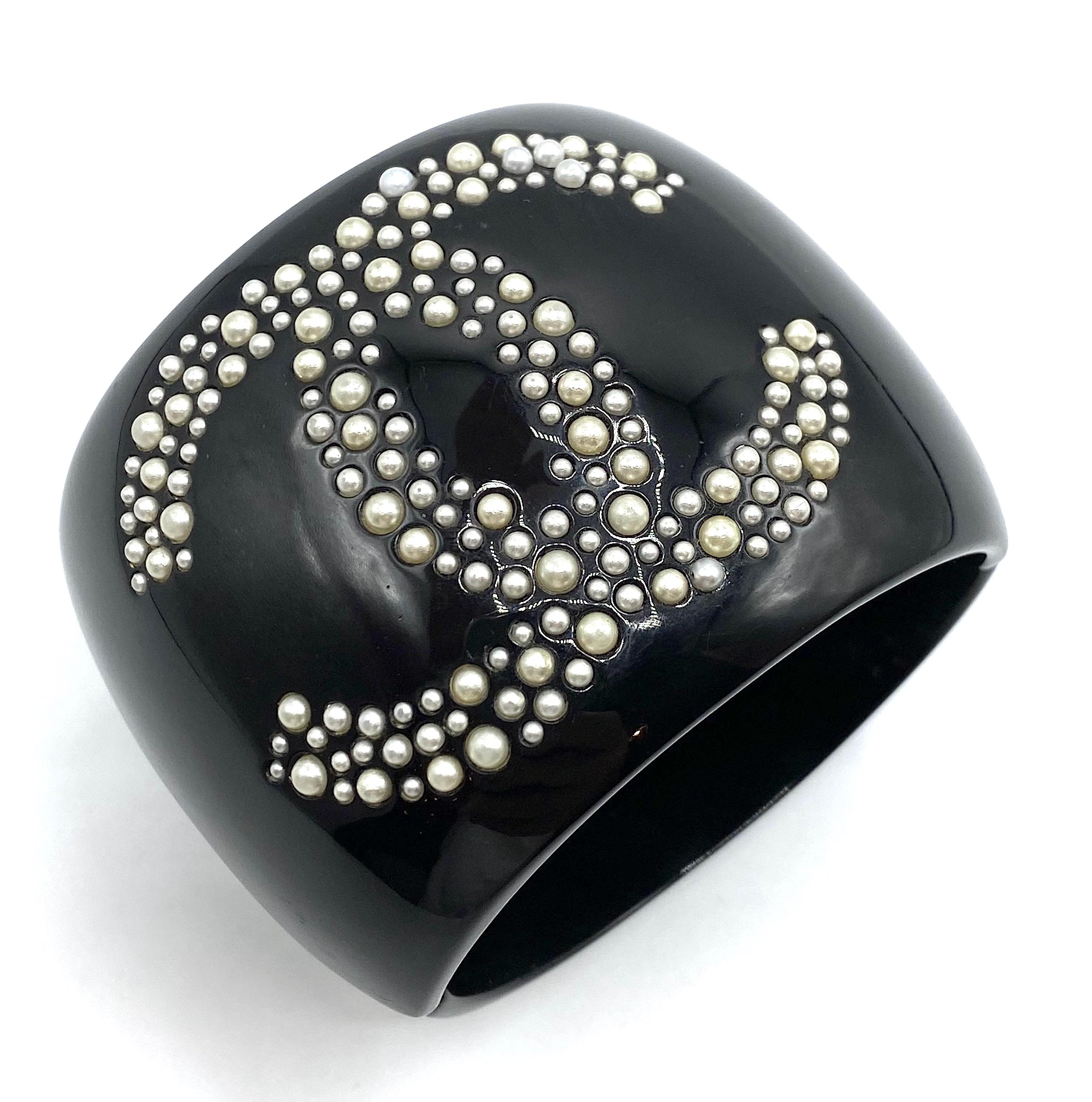 A smart looking Chanel black resin cuff style bracelet with large iconic Chanel interlocking CC logo in small and mini faux pearls. The cuff has a rhodium hinge with oval signature plaque mounted on the inside. The plaque is stamped with copyright
