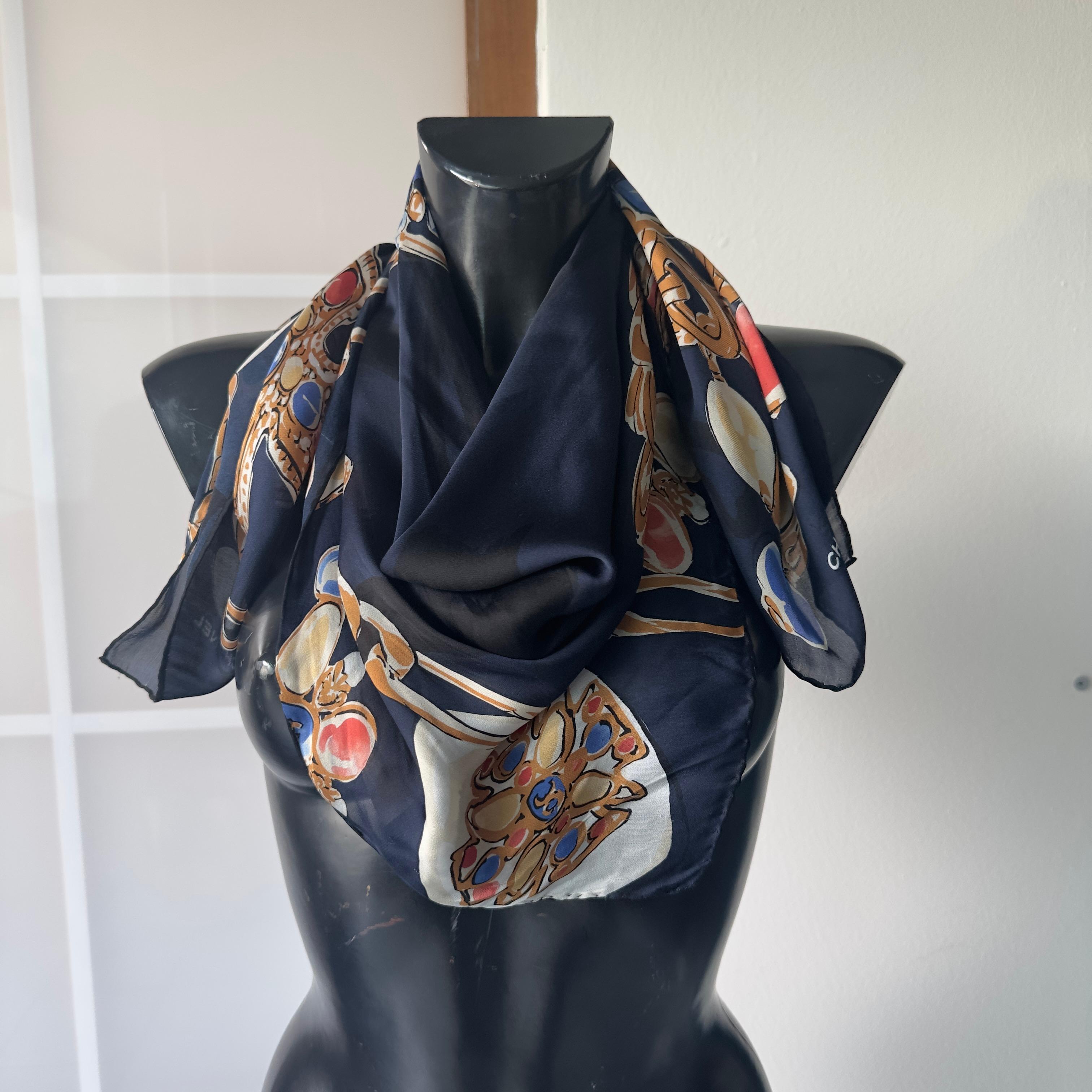 Un raro pañuelo Chanel de pura seda, adornado con un extraordinario estampado barroco con cadenas doradas, medallones enjoyados y el icónico logotipo CC en primer plano. El fondo azul noche realza el brillo de las piedras estilizadas en tonos rubí,