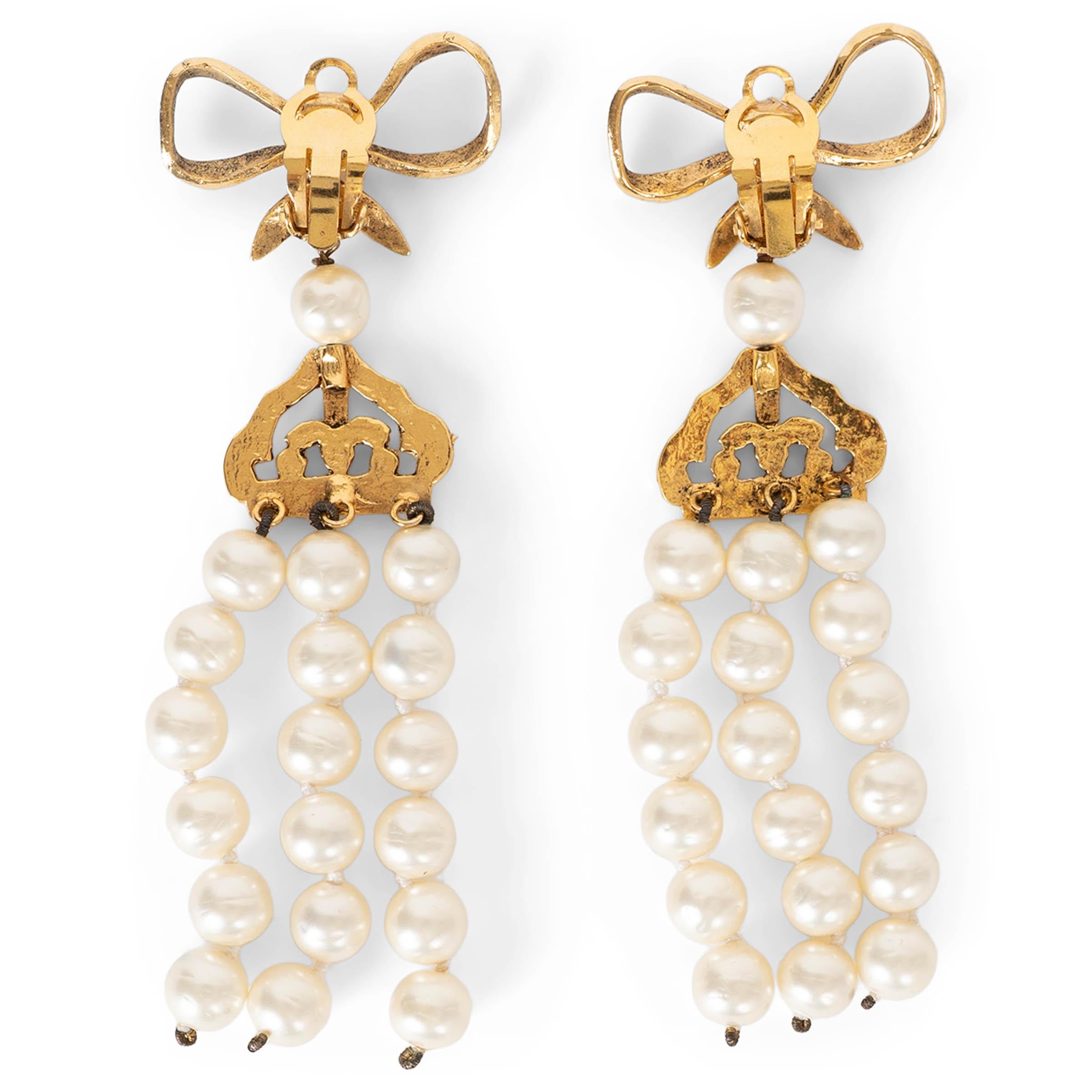 These 100% authentic Chanel chandelier clip-on earrings feature an ornate gold-tone metal bow from which a sculpted crown motif elegantly suspends three cascading strands of pearly white imitation pearls. A beautifully dramatic design that captures