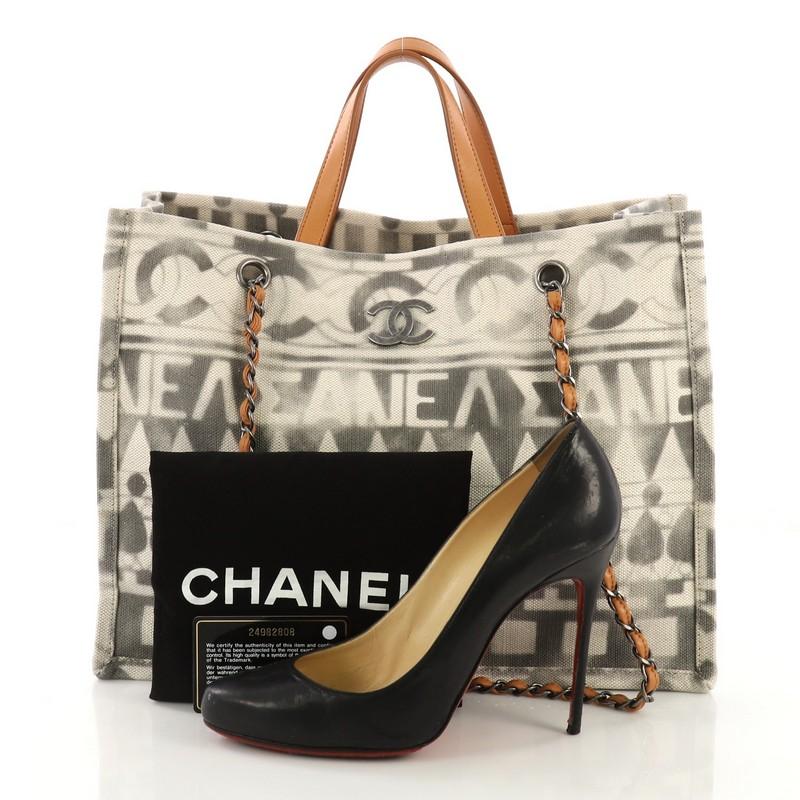 This Chanel Iliad Shopping Tote Printed Canvas Medium, crafted from gray printed canvas, features dual top leather handles, woven-in leather chain straps, and aged silver-tone hardware. Its magnetic snap button closure opens to a beige fabric