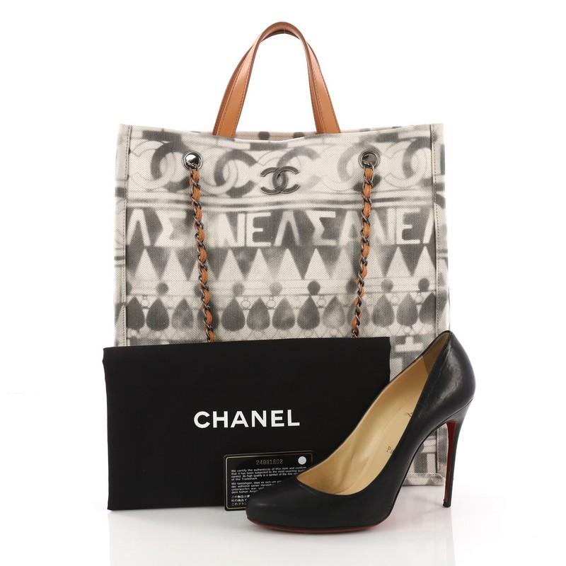 This Chanel Iliad Shopping Tote Printed Canvas Tall, crafted from off-white printed canvas, features dual top leather handles, woven in leather chain straps, and aged silver-tone hardware. Its magnetic snap button closure opens to a beige canvas