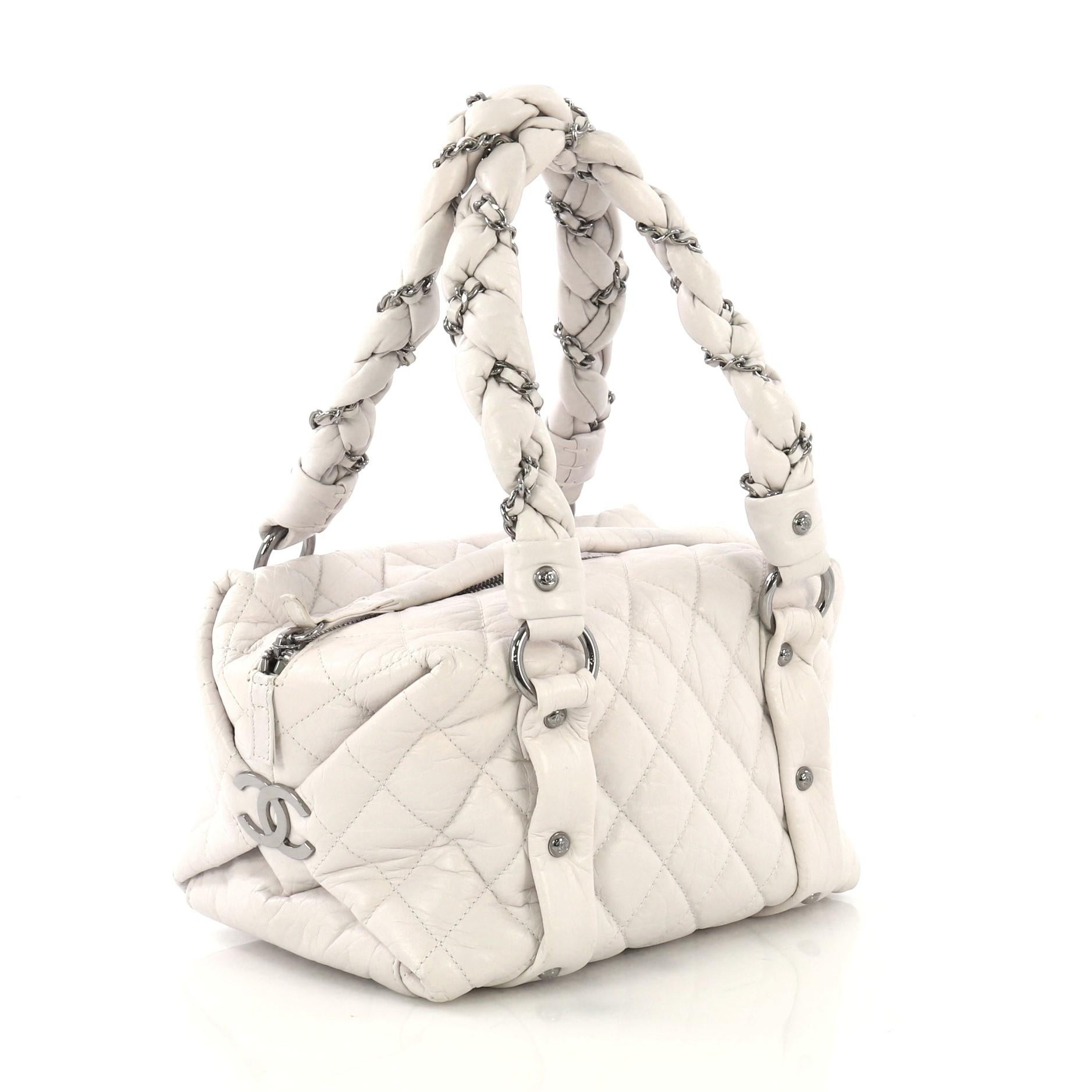 This Chanel Lady Braid Bowler Bag Quilted Distressed Lambskin Small, crafted from light grey quilted distressed leather, features intertwined woven leather chain straps, side CC logo details, and silver-tone hardware. Its top zip closure opens to a