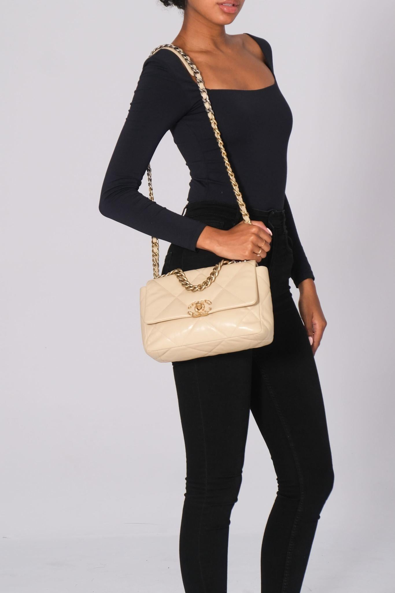 The Chanel 19 Flap Bag, made in 2019, is constructed of quilted lambskin leather in beige. It features aged gold and ruthenium hardware, a chain handle, a leather-woven chain shoulder strap, and a back slip pocket. The bag is secured with a flap and