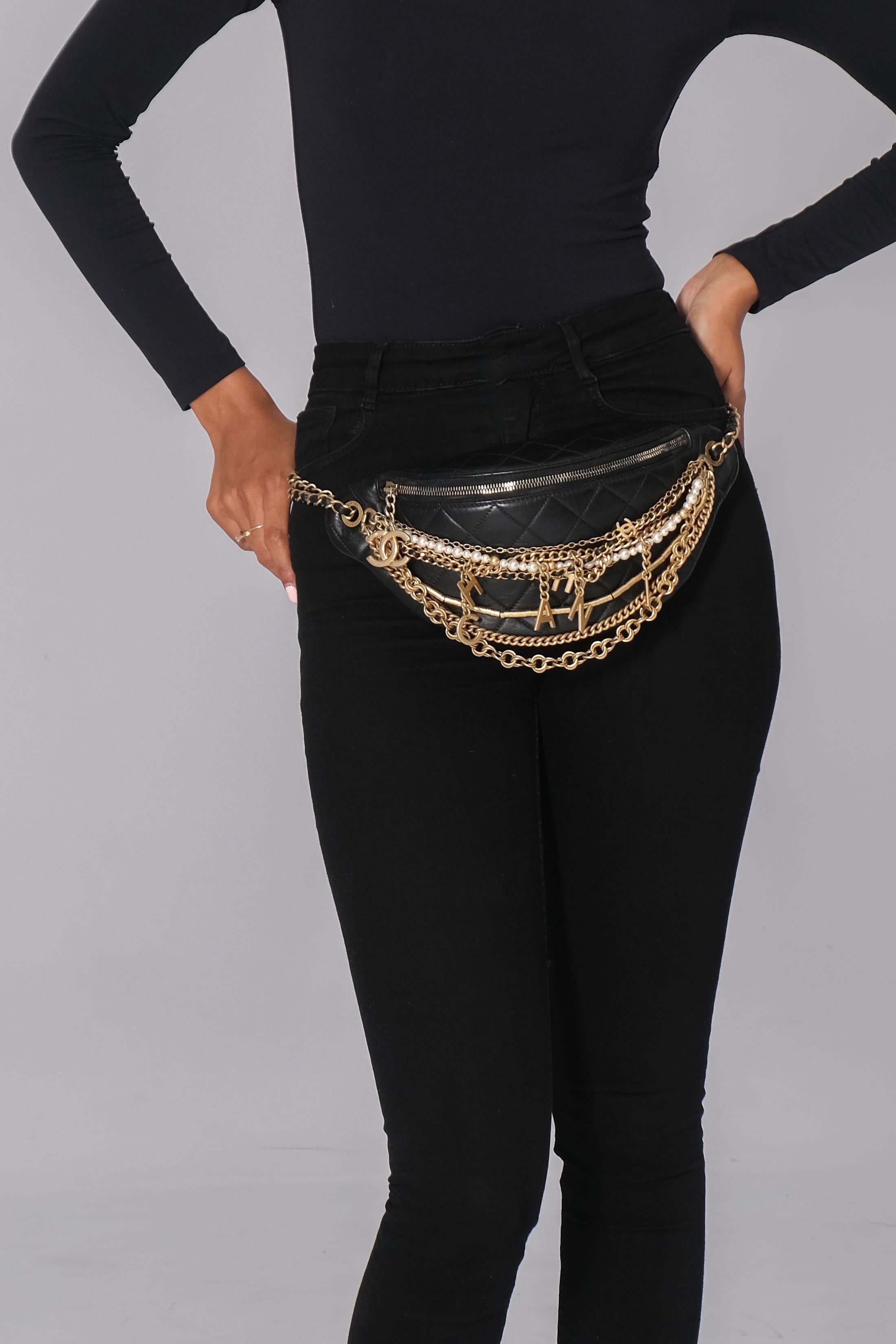 Ce sac de taille est réalisé en cuir d'agneau noir matelassé de diamants, avec des chaînes dorées à plusieurs brins suspendues à l'avant avec des charmes de logo. Le sac est doté d'une chaîne en cuir à maillons dorés et d'une sangle de ceinture