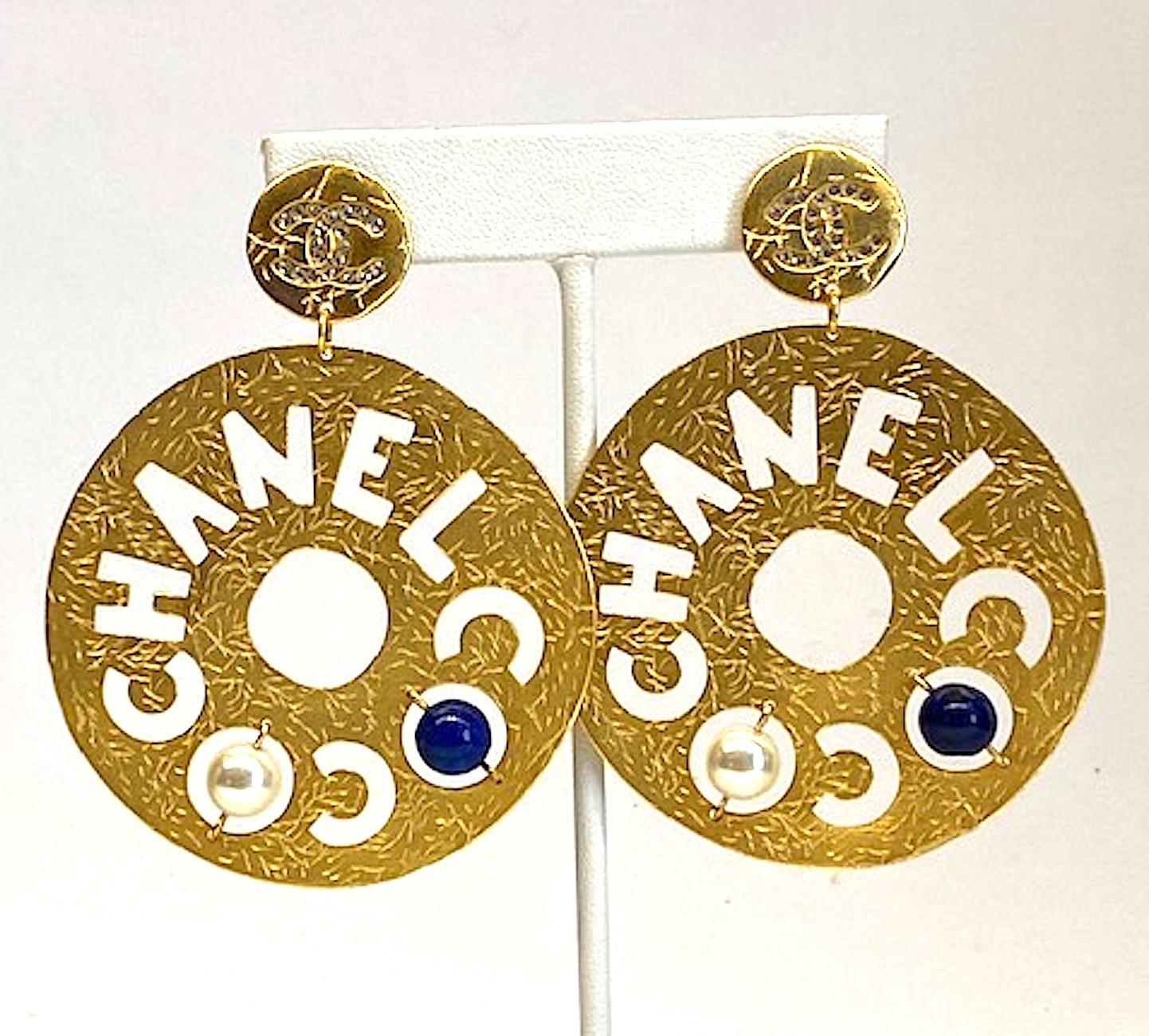 Chanel large slightly irregular round shape disk pendant Pierced earrings. They are in a satin textured gold tone and from the 2019 Autumn collection. The words Coco and Chanel are cut out from the disk. The two Os in Coco have a faux lapis glass