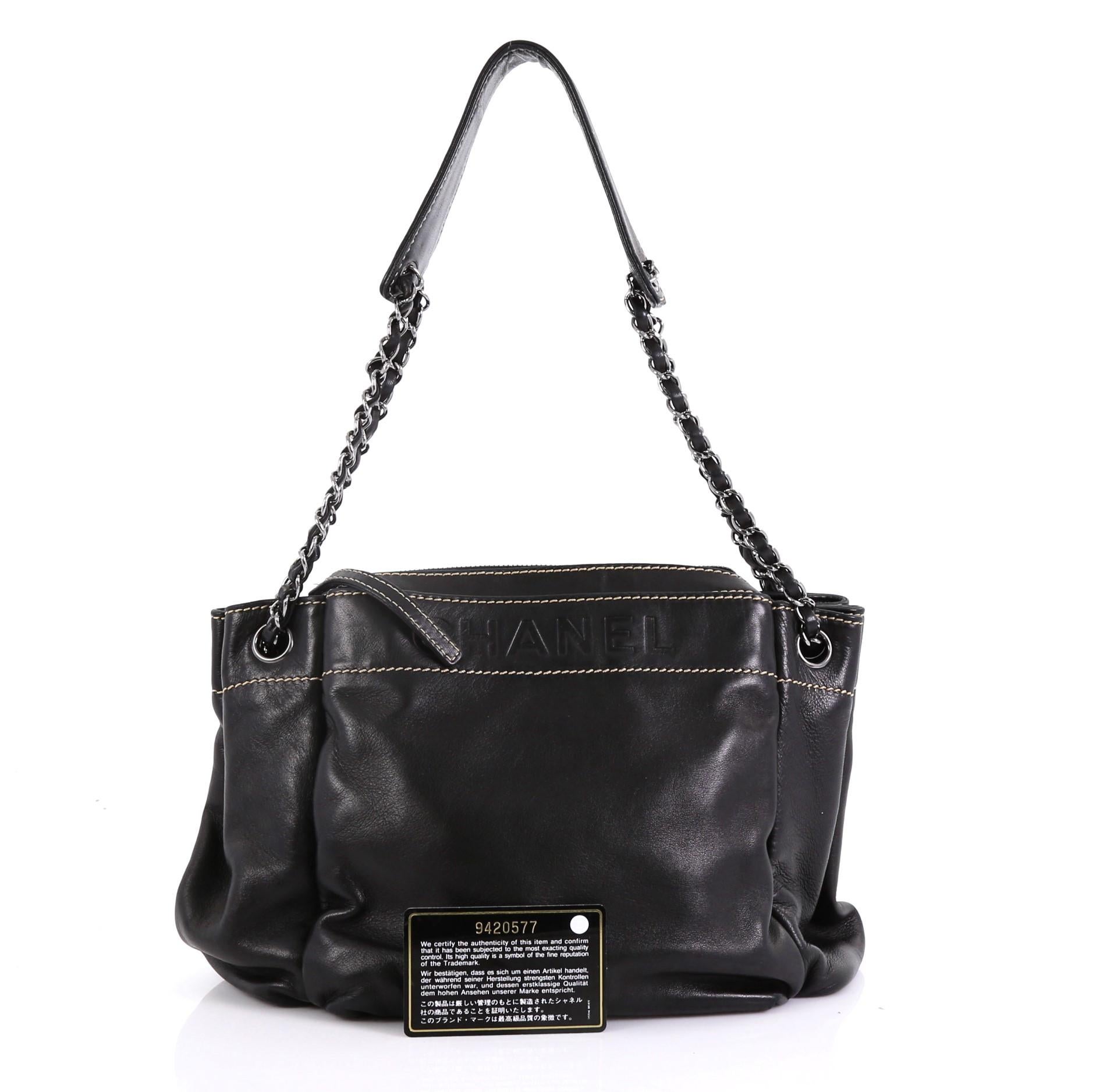 This Chanel Lax Accordion Tote Leather Medium, crafted from black leather, features woven-in leather chain straps with leather pad, embossed logo at the front, and gunmetal-tone hardware. It opens to a gold fabric interior with zip pocket. Hologram