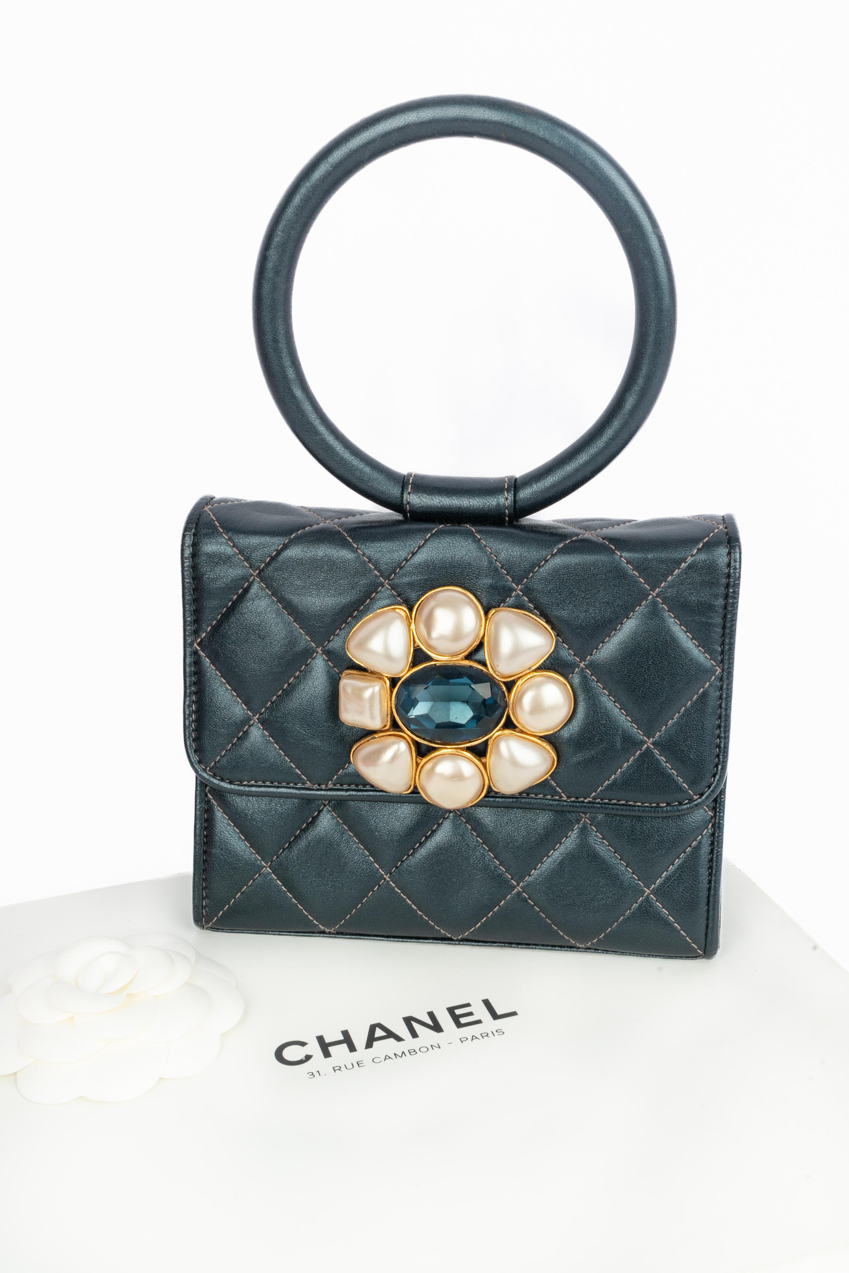 CHANEL - (Made in France) Quilted metallic-blue leather bag ornamented with a golden metal jewelry, rhinestones, and a costume pearly cabochon. Round handle. Piece with a serial number and an authenticity card. 1991-1992 Fall-Winter Collection under
