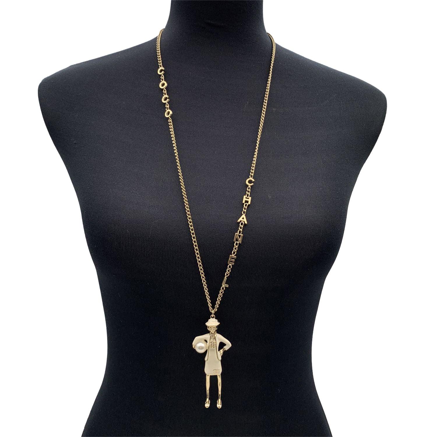 Light gold metal long chain necklace with Mademoiselle Coco pendant by CHANEL. Silver metal mademoiselle Coco figurine holding a pearl with beige enamel. C.H.A.N.E.L. letters on the chain. Lobster closure. Pendant's max height: 3 inches - 7.6 cm.