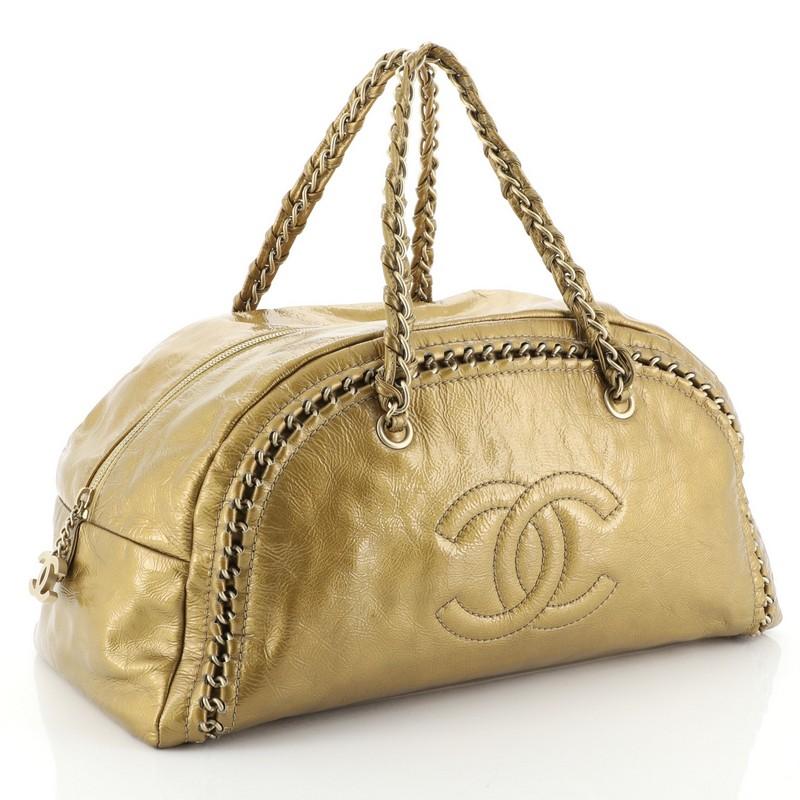 This Chanel Luxe Ligne Bowler Bag Patent Large, crafted from gold patent leather, features braided leather and chain straps, embedded chain around trim, frontal stitched CC logo, and matte gold-tone hardware. Its zip closure opens to a gold leather