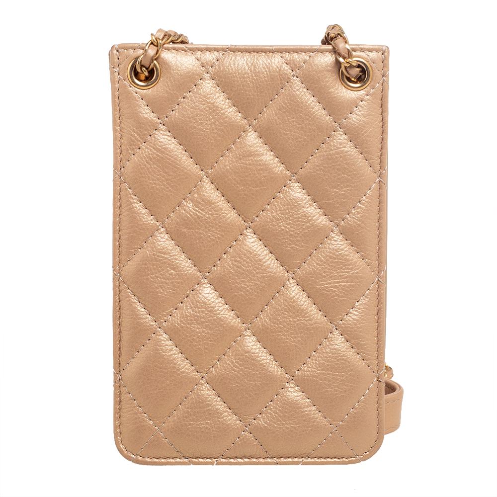 A Chanel phone holder for an opulent touch to the daily OOTD. It is made from metallic beige quilted leather, with a CC logo adorning the front. It has a sturdy woven chain strap and fabric-lined compartments to house your phone, cards, and