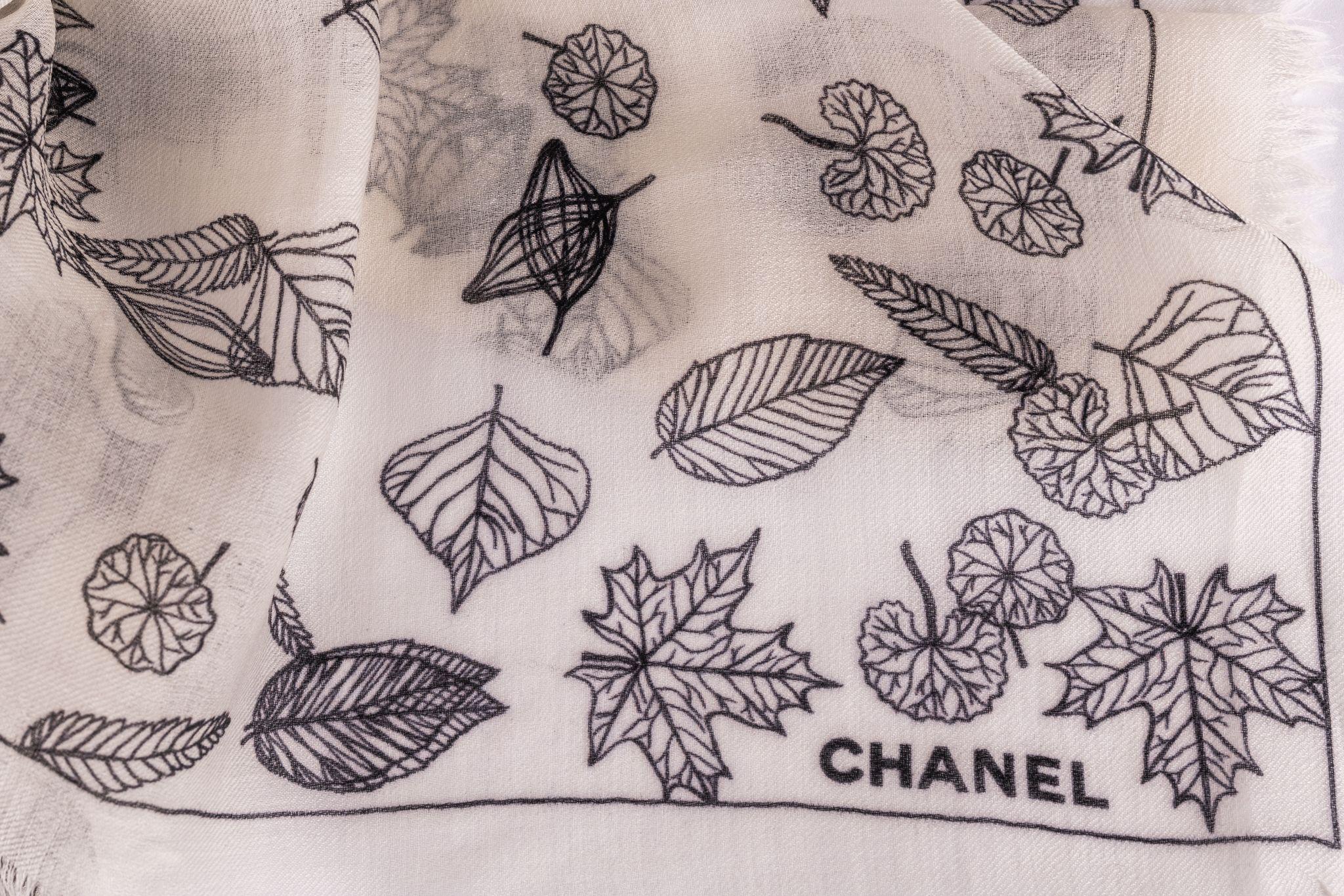 Scialle in cashmere Chanel di colore bianco con un grande logo CC al centro. Il logo è disegnato con delle foglie, così come la cornice della sciarpa. L'articolo è in condizioni nuove.