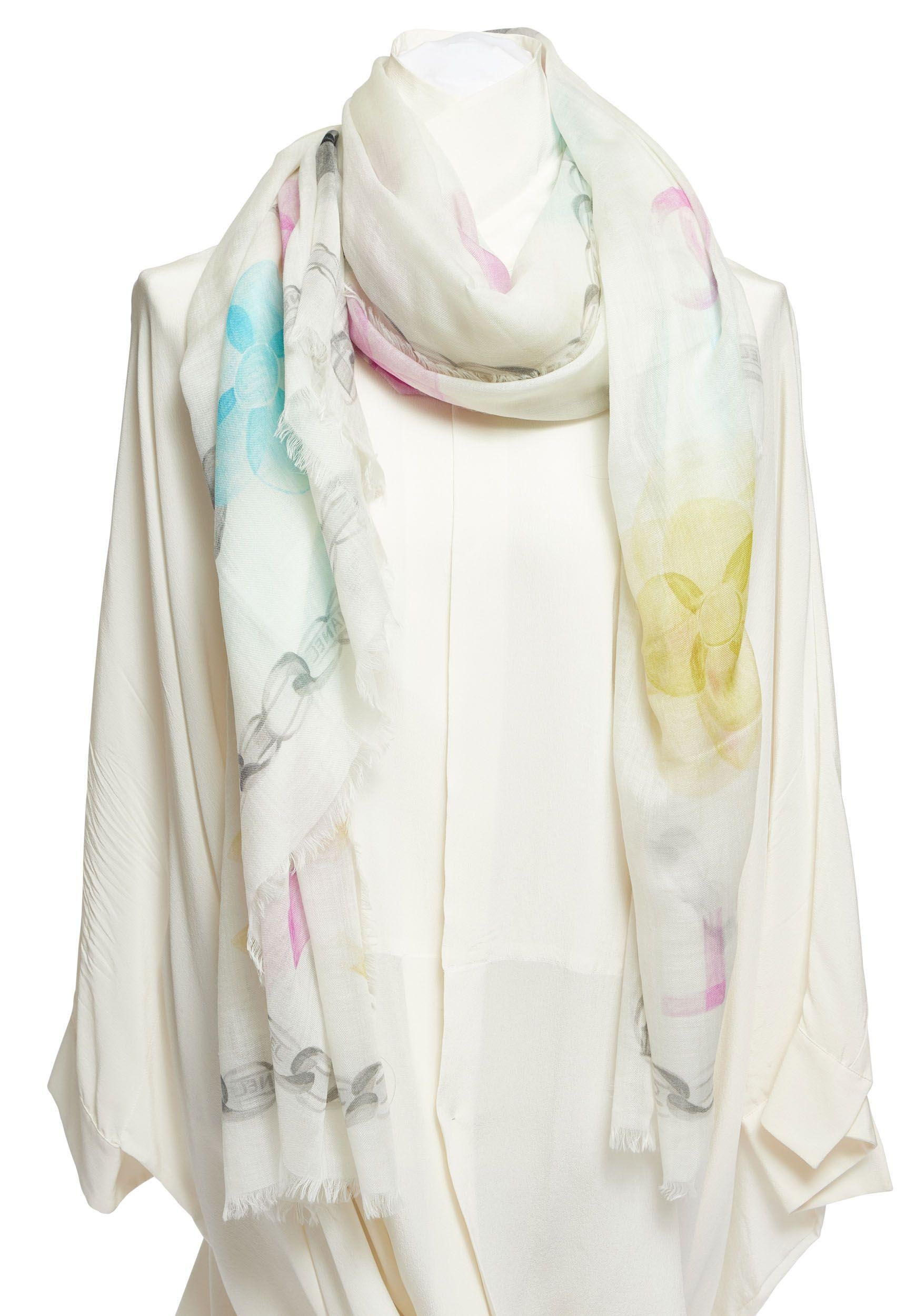 Chanel white cashmere white shawl with multicolor camellias on it and a black trim out of flowers and CC logos. The edges of the scarf are fringed. It's a big piece which can be worn as a cape or just as scarf since it's so light and soft. It is