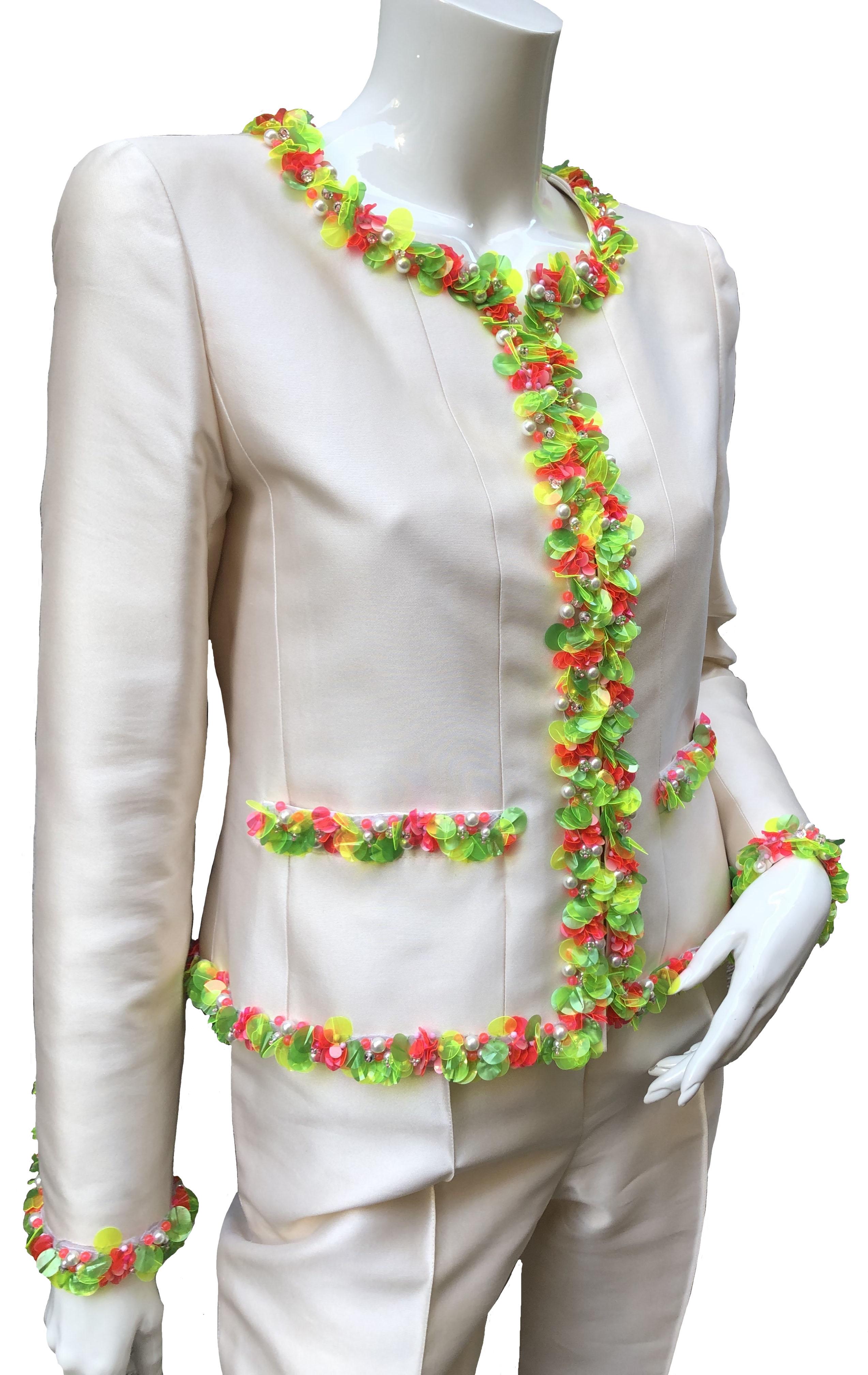 This pre-owned off-white silk pant suit from Chanel features large green and coral sequins piping. 
A short jacket with long sleeves, with a hook and loop fastener system on the front, two patch pockets, straight pants, zipped on the side,
