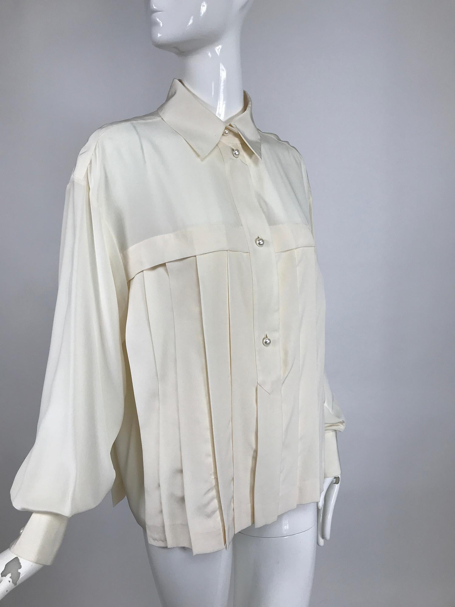 Blusa Chanel de seda blanca plisada de manga larga, con sorprendentes botones perlados con el logotipo.La blusa tiene una tapeta delantera con cuello de ala, pliegues verticales bajo el canesú por delante y por detrás. Las mangas largas son enteras,