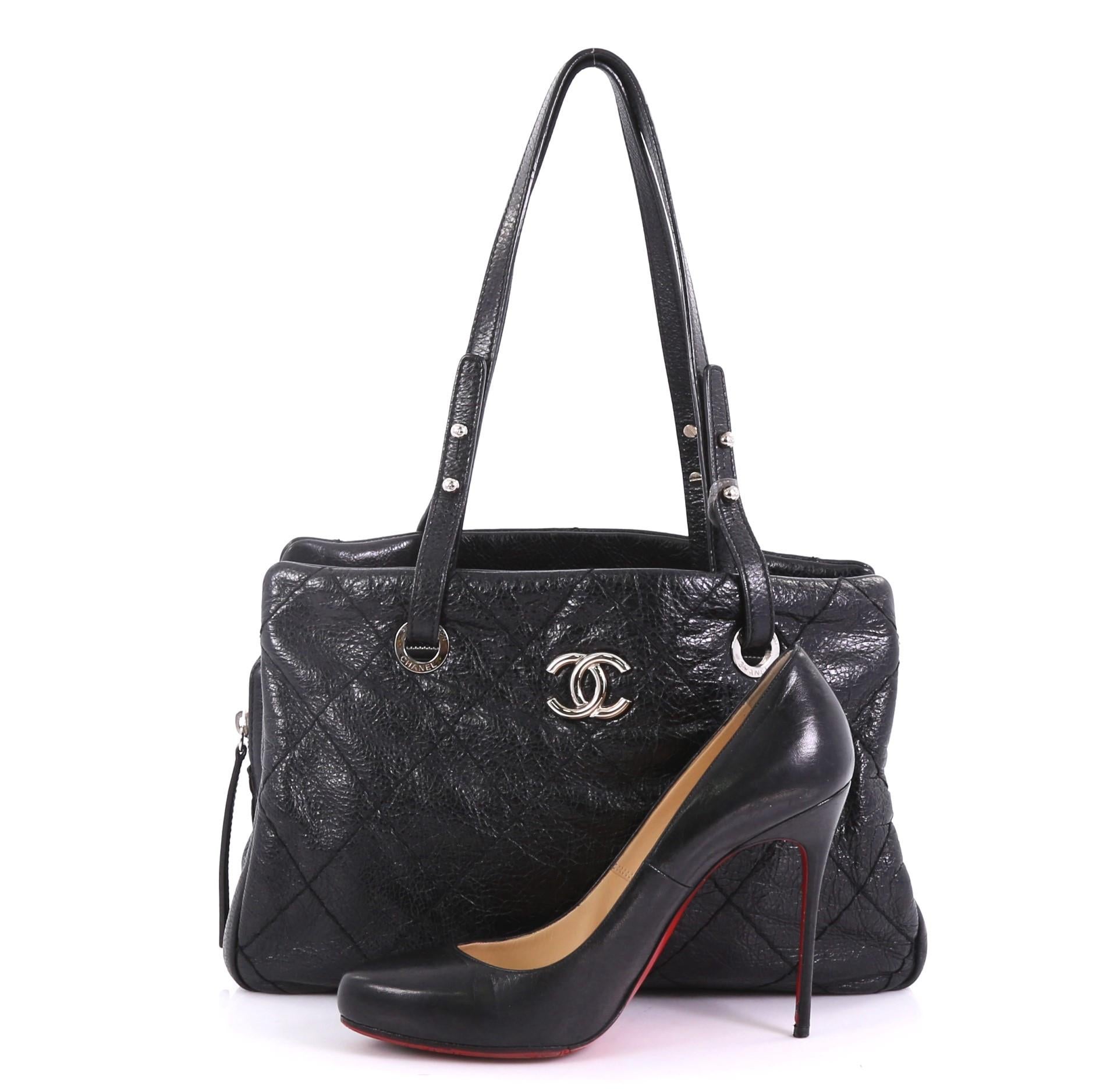 This Chanel On The Road Shopping Tote Quilted Leather Medium, crafted from black quilted leather, features dual flat leather handles, protective base studs, and silver-tone hardware. It opens to a black satin interior with a middle zip compartment