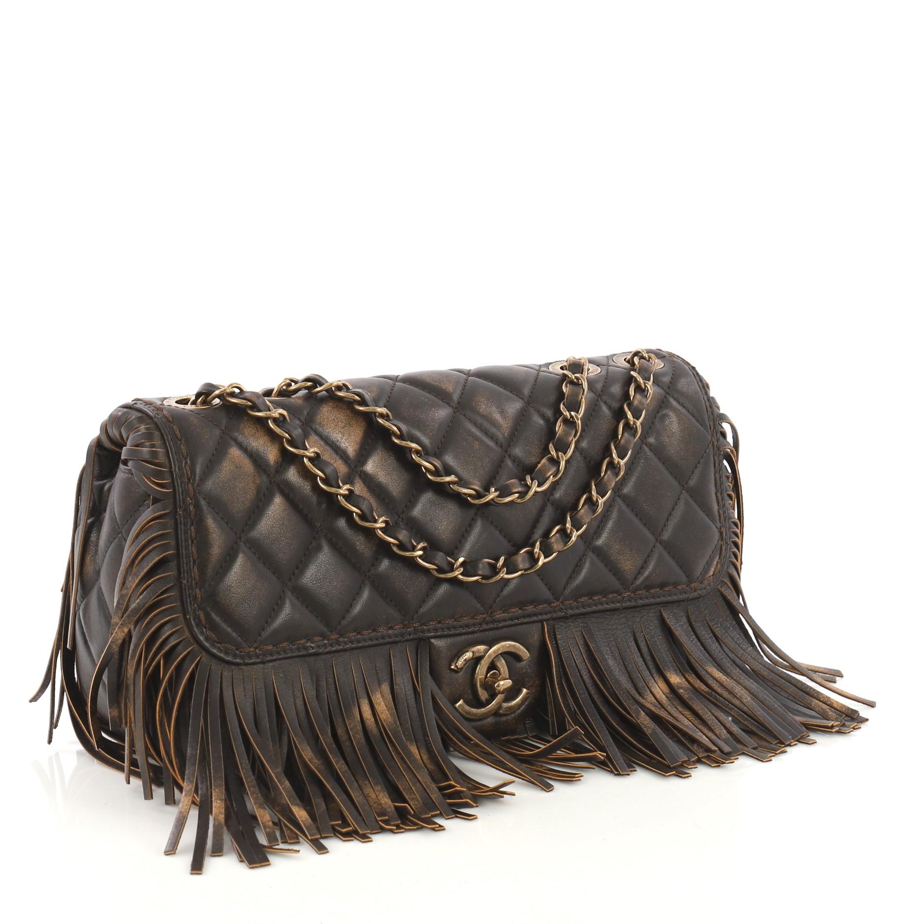 This Chanel Paris-Dallas Fringe Flap Bag Quilted Leather, crafted from brown quilted leather, features woven-in leather chain straps, frontal flap, fringe detailing, and aged gold-tone hardware. Its CC turn-lock closure opens to a burgundy fabric