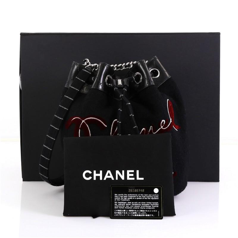 This Chanel Paris-Hamburg Drawstring Bucket Bag Embroidered Wool Small, crafted from black embroidered wool, features chain link strap with leather pad, embroidered Chanel Paris on front, and silver-tone hardware. Its drawstring closure opens to a